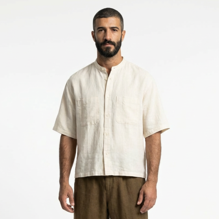A man with a beard wearing a cream-colored short-sleeve shirt and brown pants standing against a plain white background.