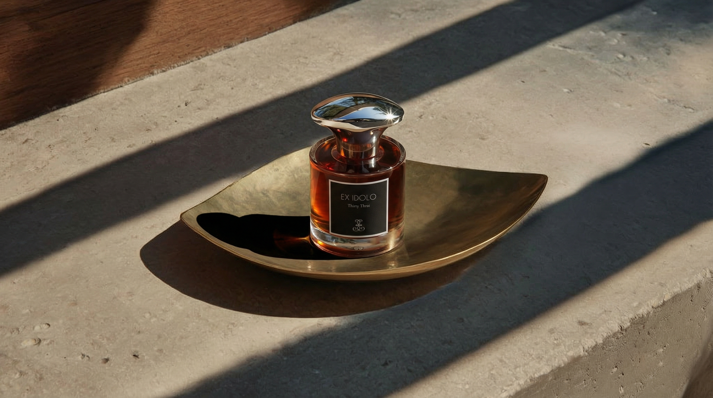 A glass perfume bottle labeled 'EX IDOLO Thirty Three' on a gold-colored tray in sunlight with shadows.