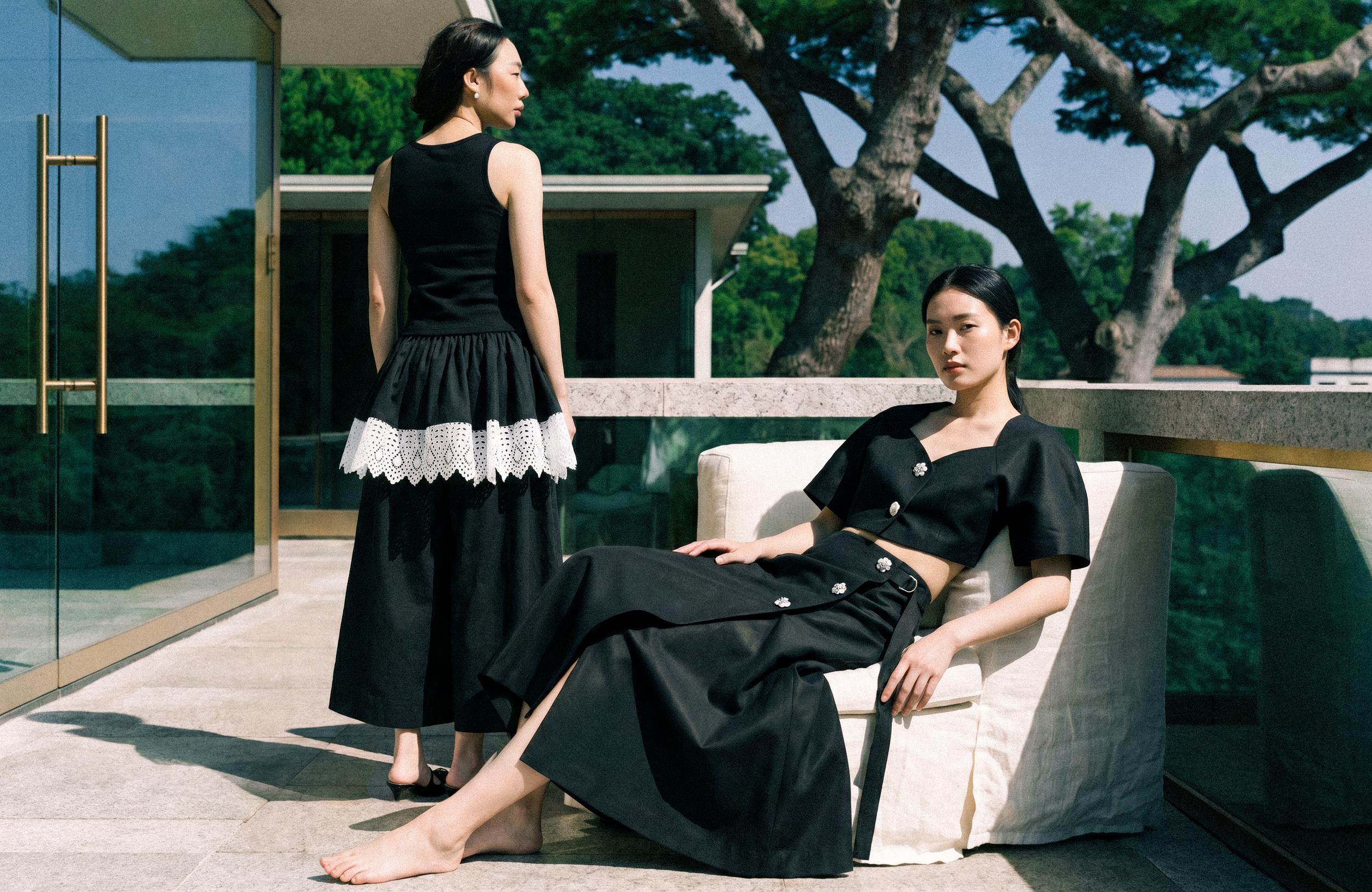 Two women in fashionable black outfits outside on a patio, one sitting on a white chair with her legs crossed and the other standing beside her, with green trees and a modern building in the background.