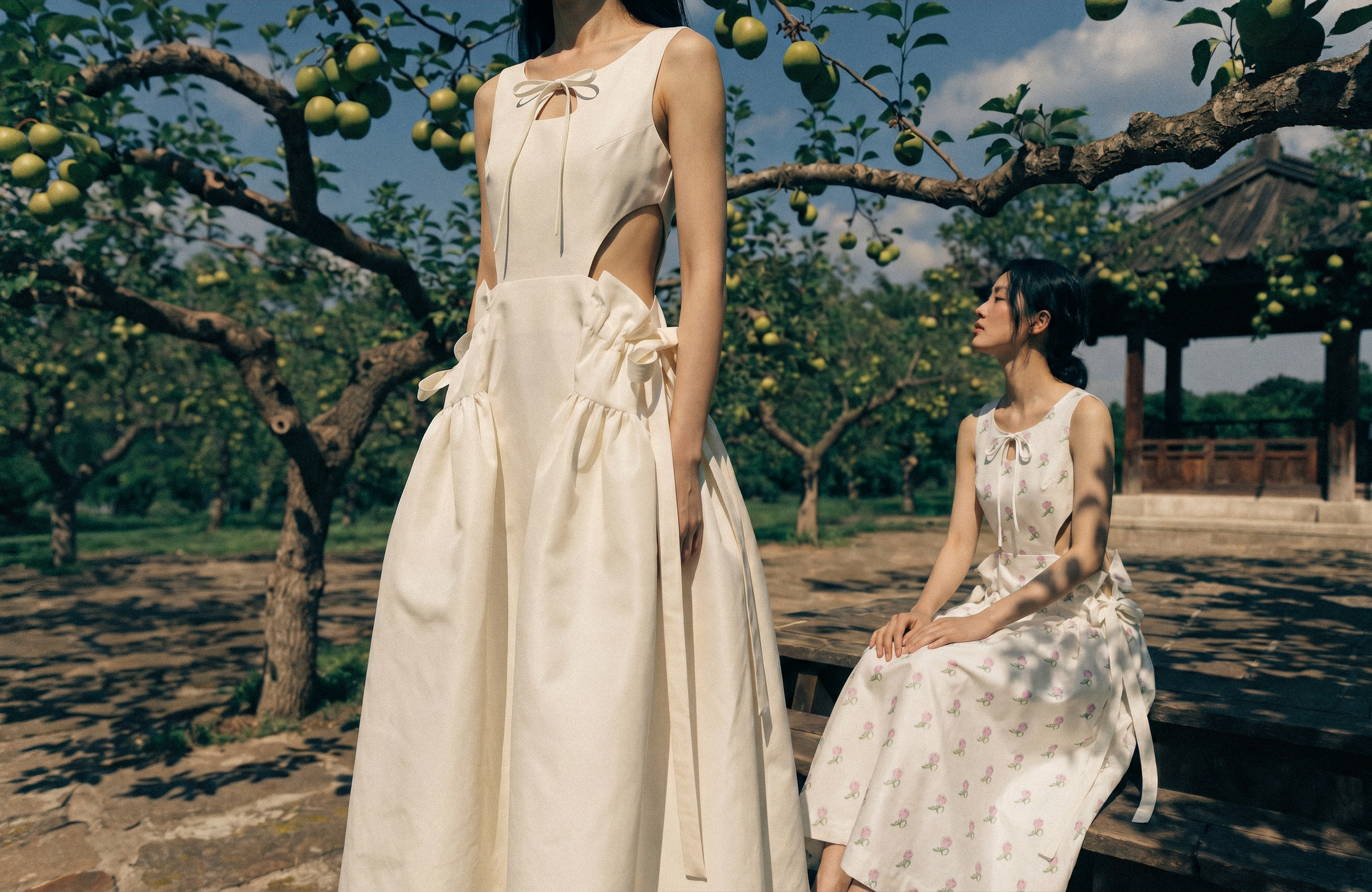 Two women in vintage-style dresses are outdoors in a garden with apple trees. One woman stands with her face not visible, wearing a cream dress with cutouts and a bow on the neckline. The other woman, sitting on a bench, has dark hair and is wearing a sleeveless dress with a cherry pattern, looking at the standing woman.