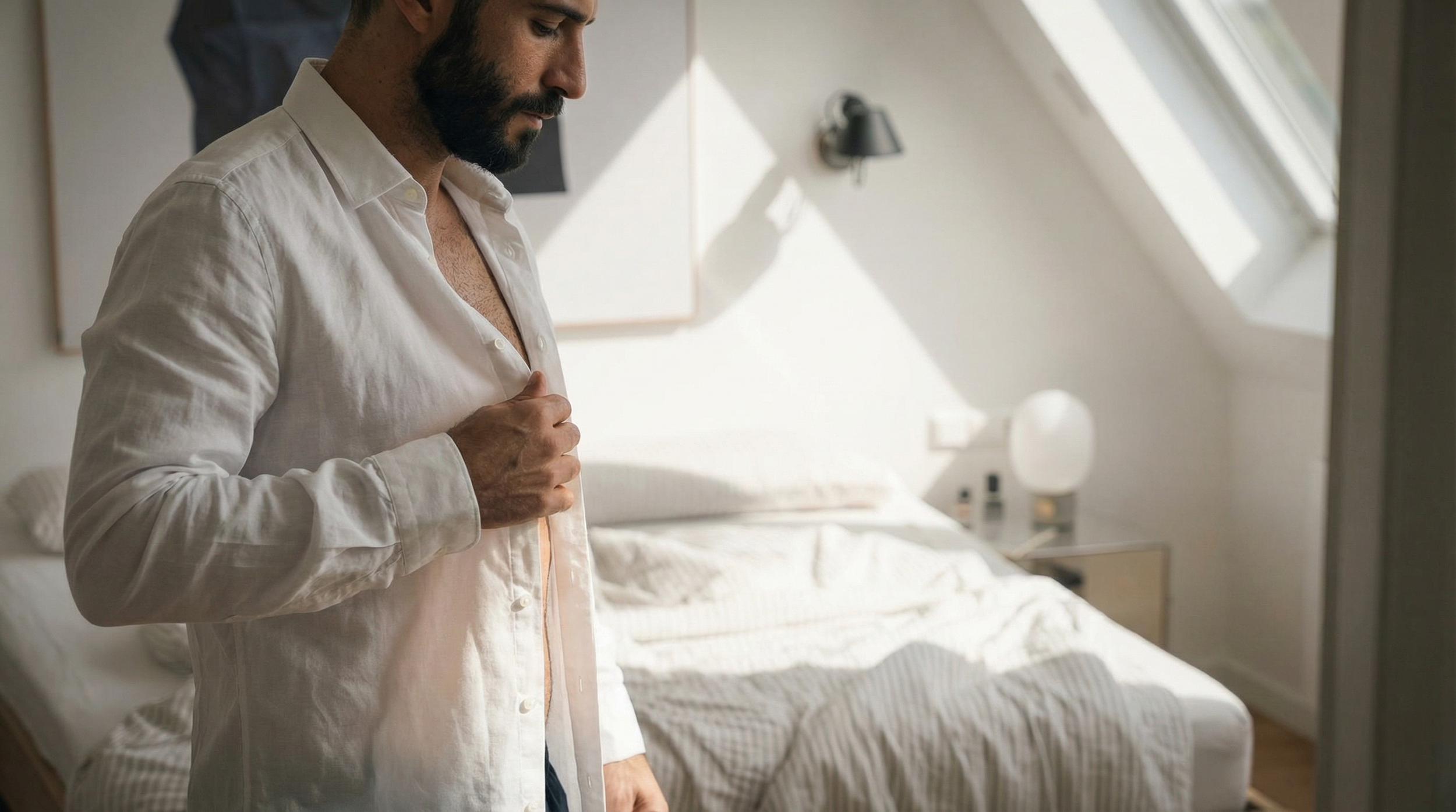 A man with a beard in a white shirt adjusting his shirt in a bright bedroom with a sloped ceiling and a bed in the background.