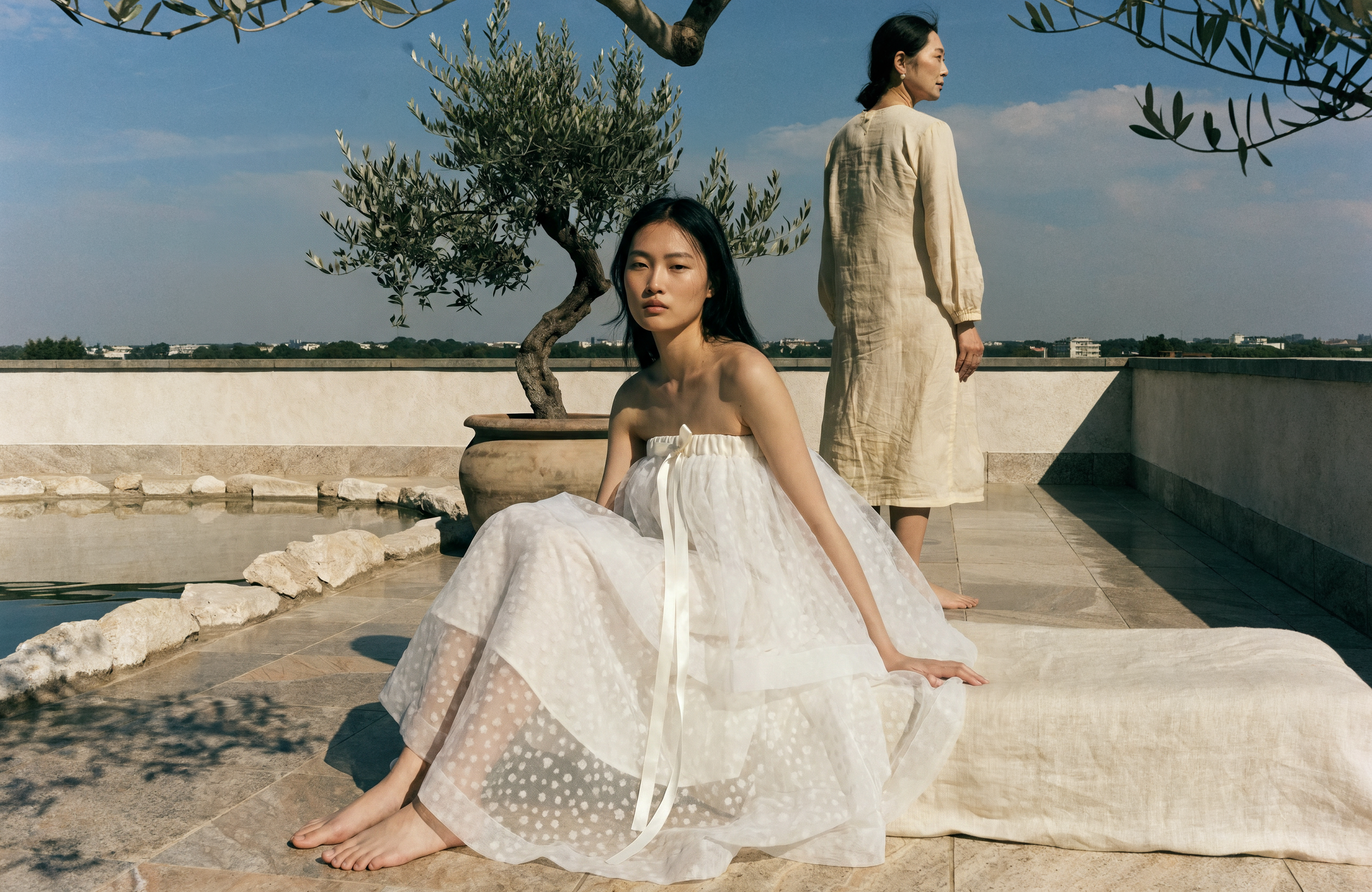 Two women with black hair and light skin on a tiled rooftop with potted trees and a cityscape in the background. One woman sits on the ground in a white strapless dress, while the other stands in a long beige dress. They are barefoot, and sunlight casts shadows on the ground.