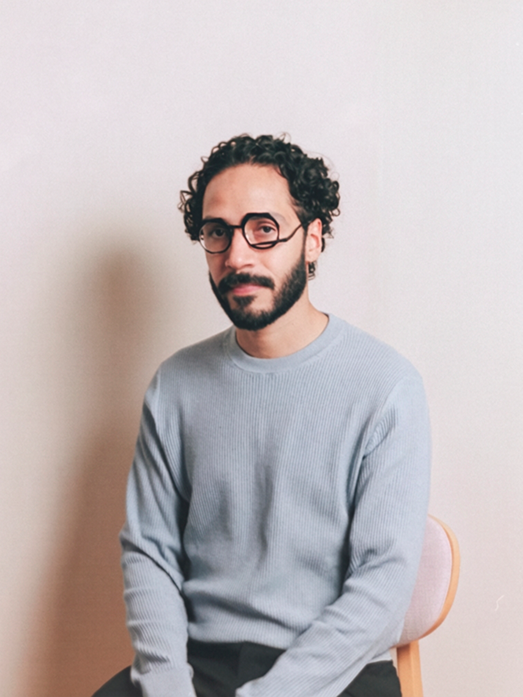A man with curly hair and glasses sitting on a wooden chair against a plain light-colored wall, wearing a light gray striped sweater.