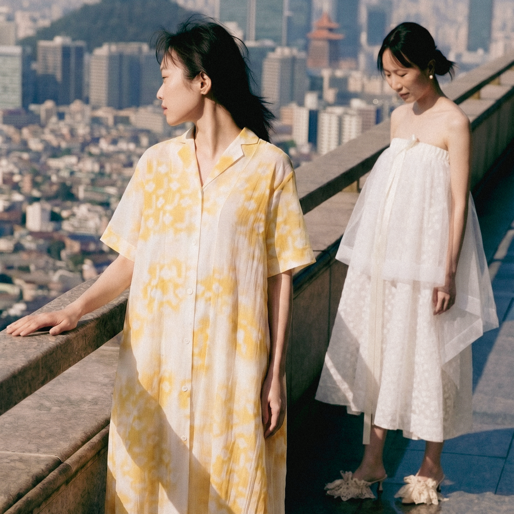 Two women are standing on a rooftop, overlooking a city skyline with tall buildings. One woman is wearing a yellow and white patterned dress, and the other woman is in a white strapless dress with lace details. The woman in the white dress is holding a phone in her hand.