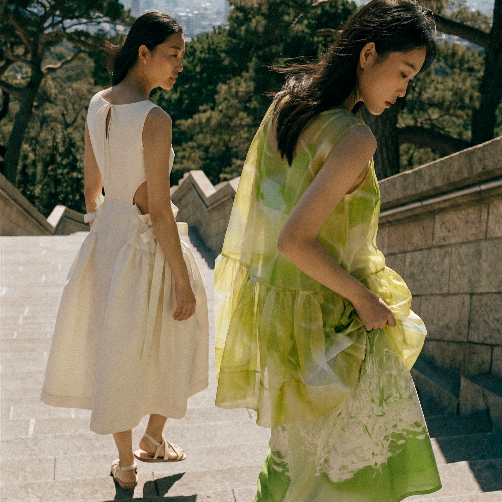 Two women walking outdoors on a sunny day, one wearing a white dress and the other wearing a yellow and green sheer dress, with trees and a stone wall in the background.