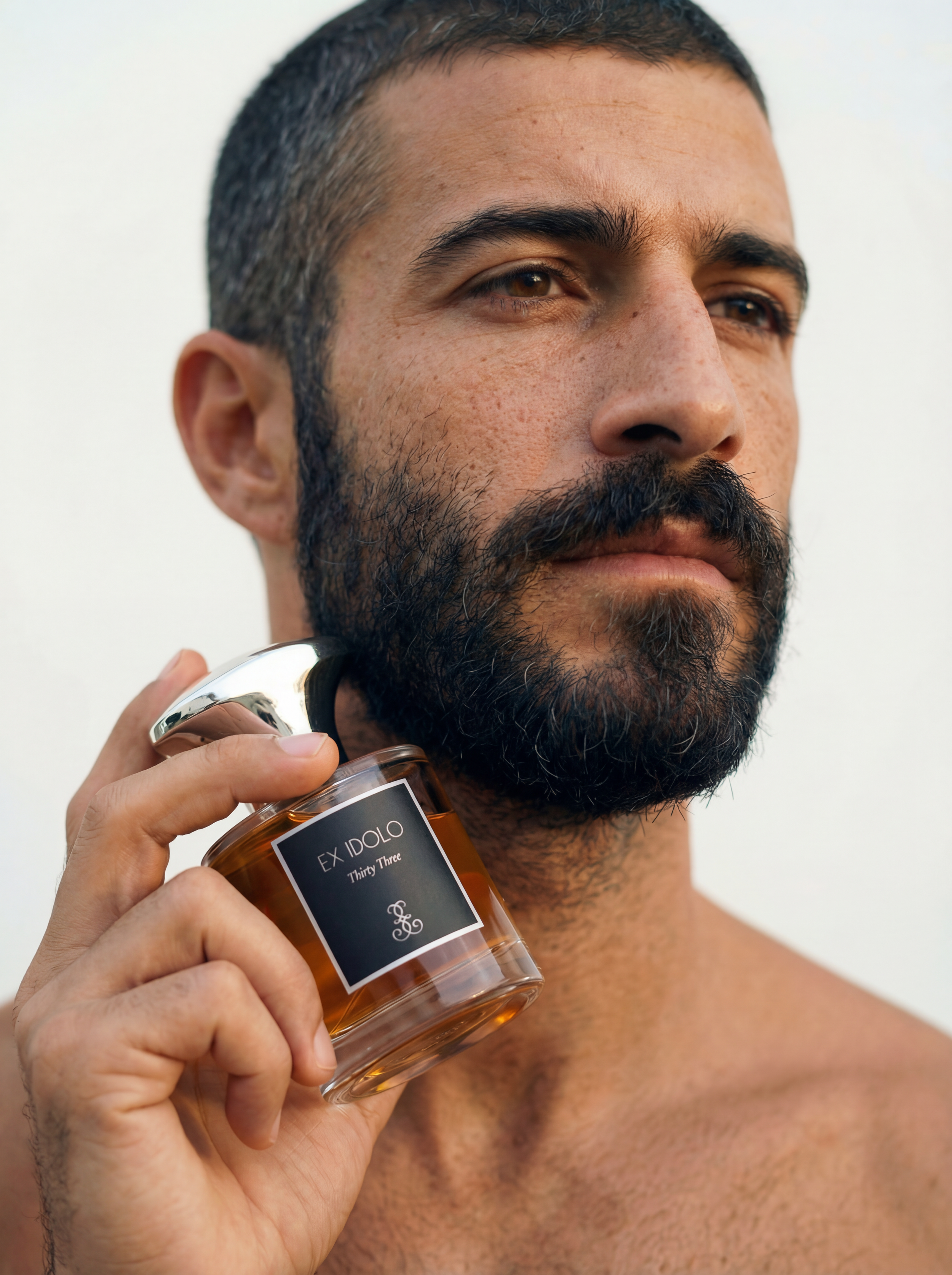 Close-up of a man with a beard holding a jar labeled 'Ex Idolo Thirty Three' near his face with a neutral background.