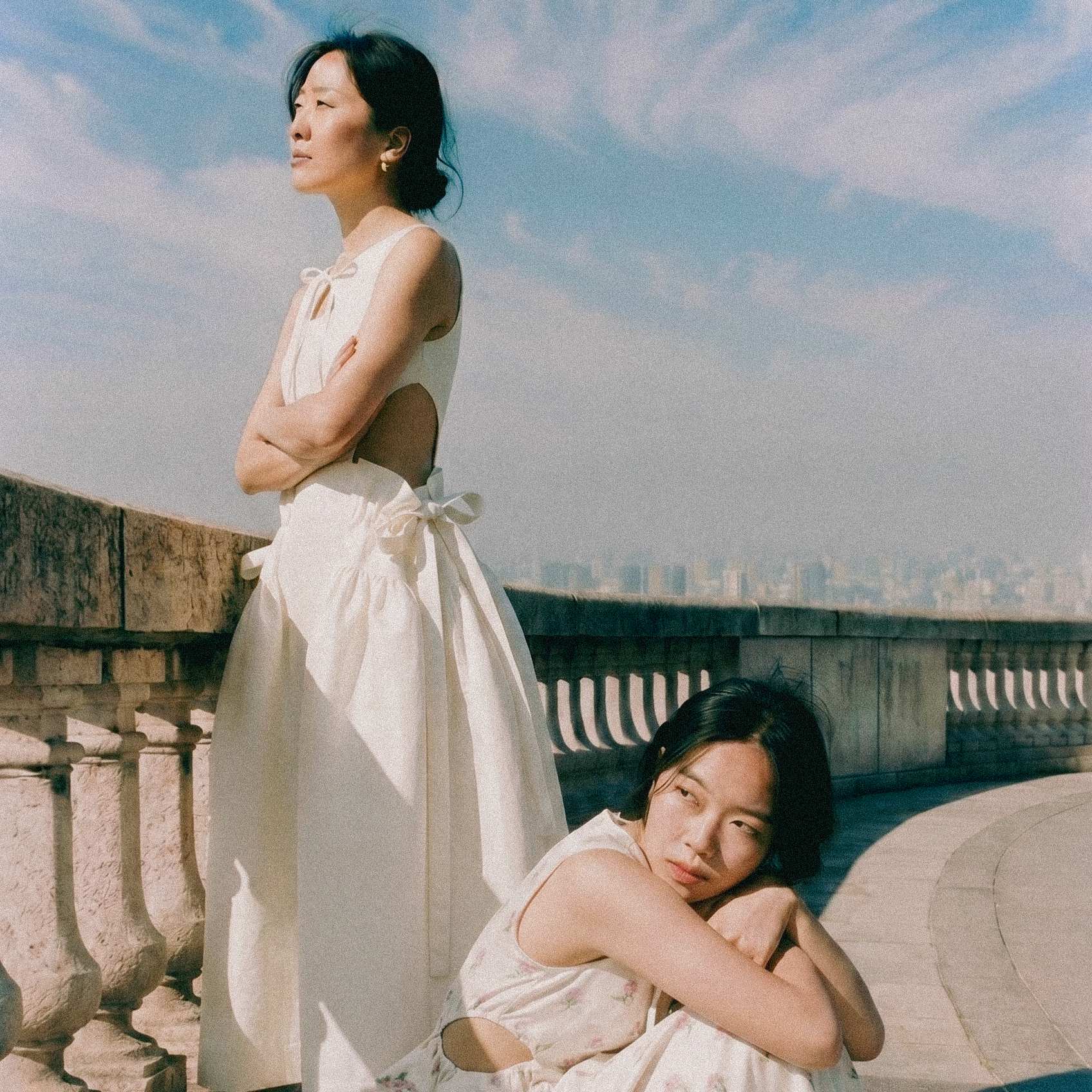 Two women on a rooftop, one standing and the other sitting, with a city skyline and blue sky in the background.