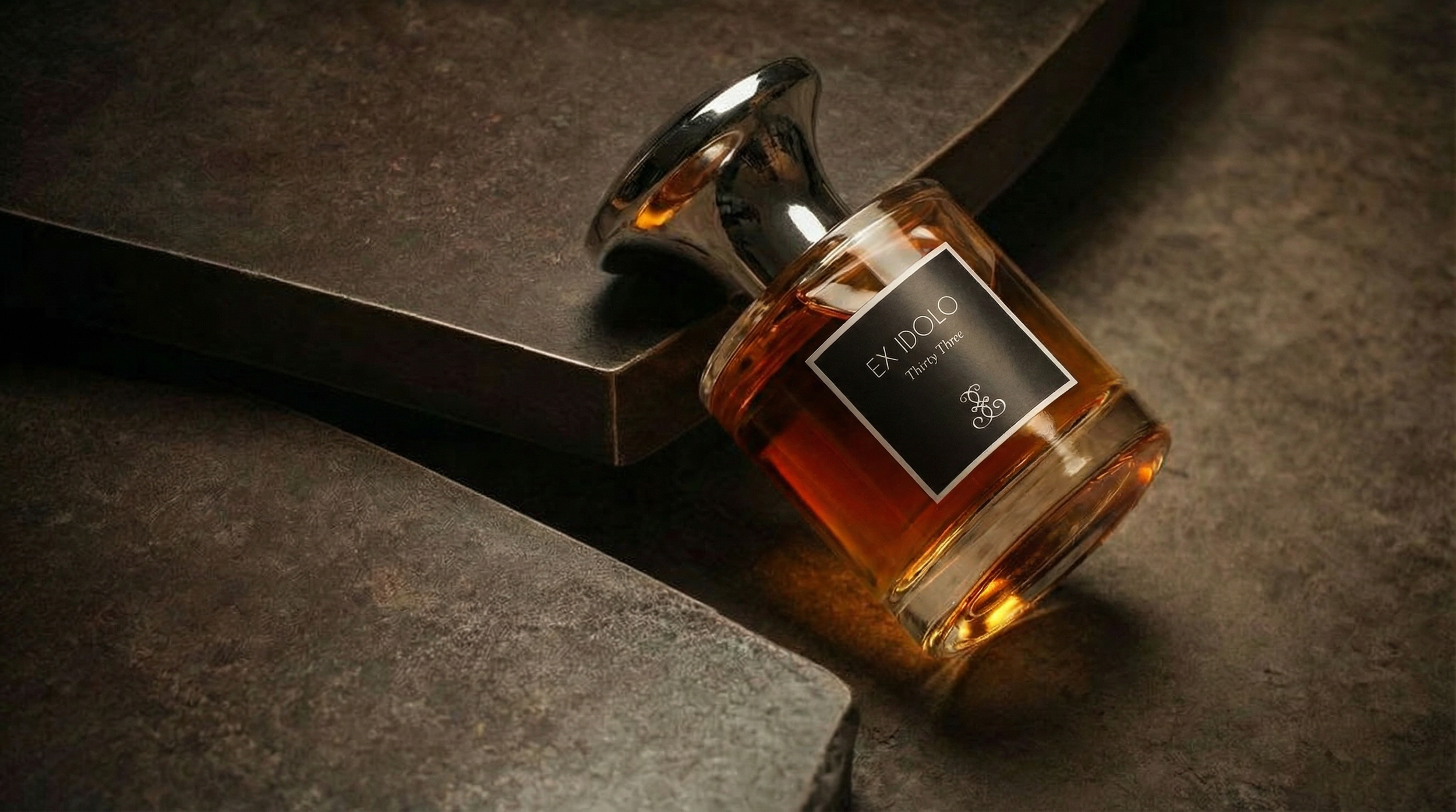 A bottle of perfume labeled 'EX IDOLO Thirst Three' lying on a textured dark surface near a dark-colored box.
