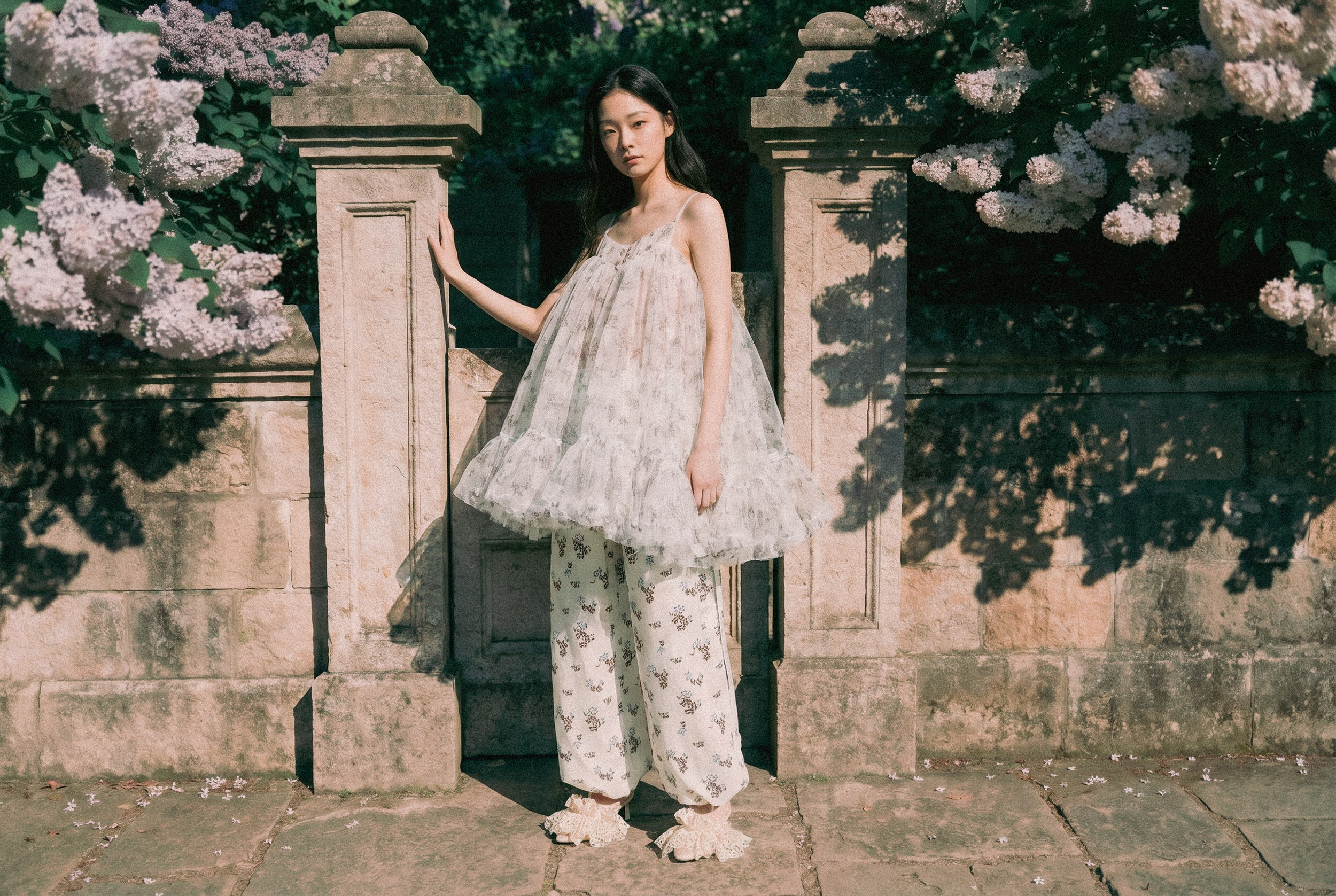 A young woman in a flowy, floral dress and lace shoes standing outdoors next to a stone wall with flowering bushes, dappled sunlight, and shadows.