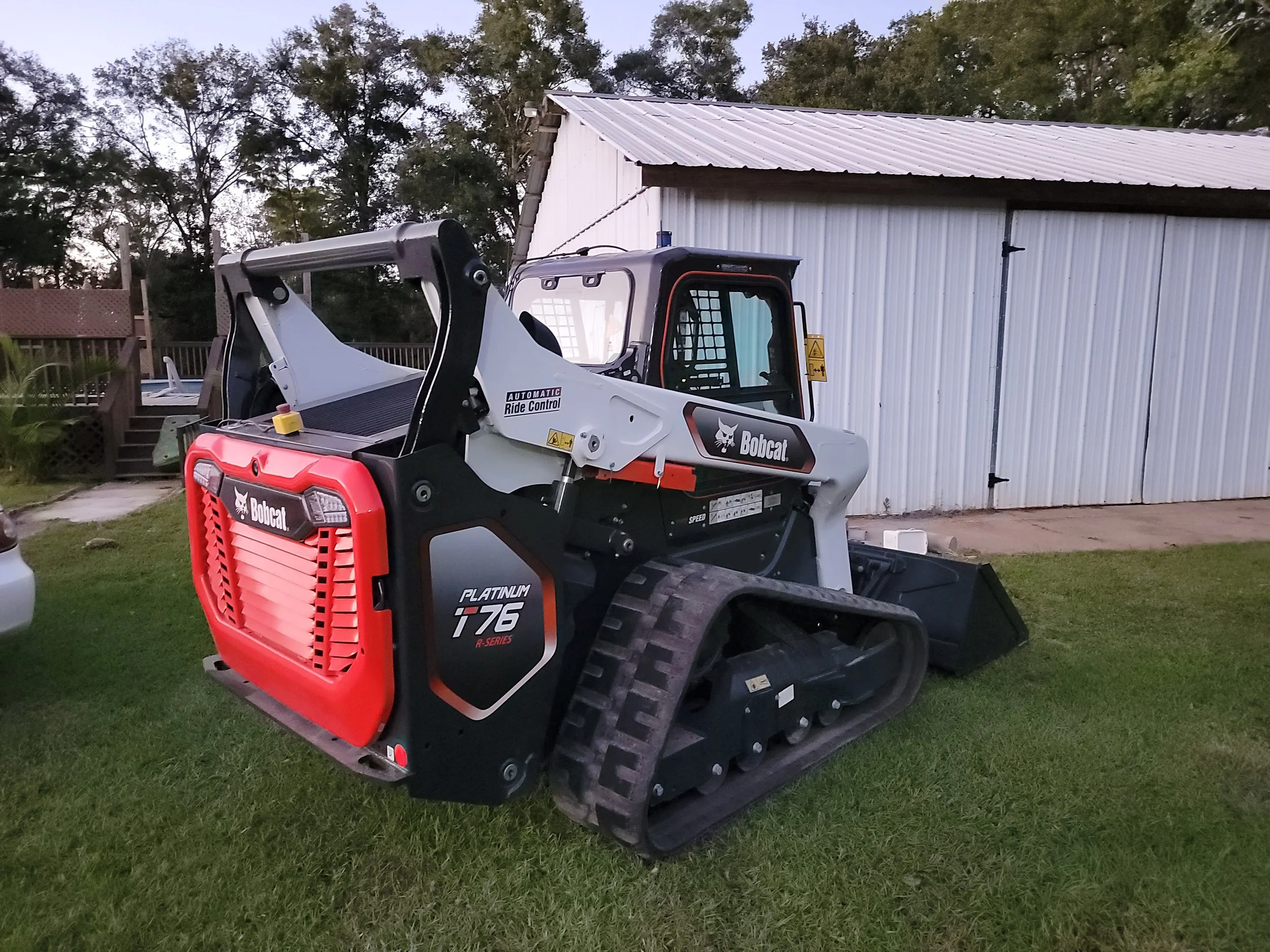 A Bobcat compact tracked loader parked on a grassy lawn in front of a white metal building with a sloped roof, trees in the background, during dusk.