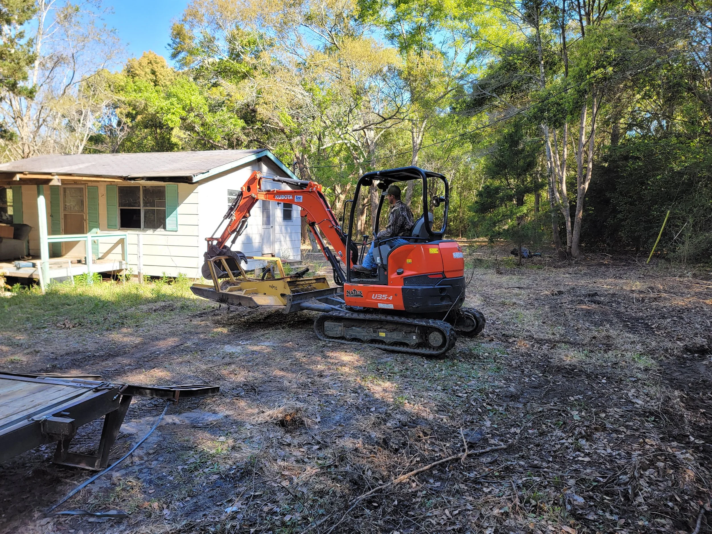 A person operating a small orange excavator in a backyard with a white house and green trees.