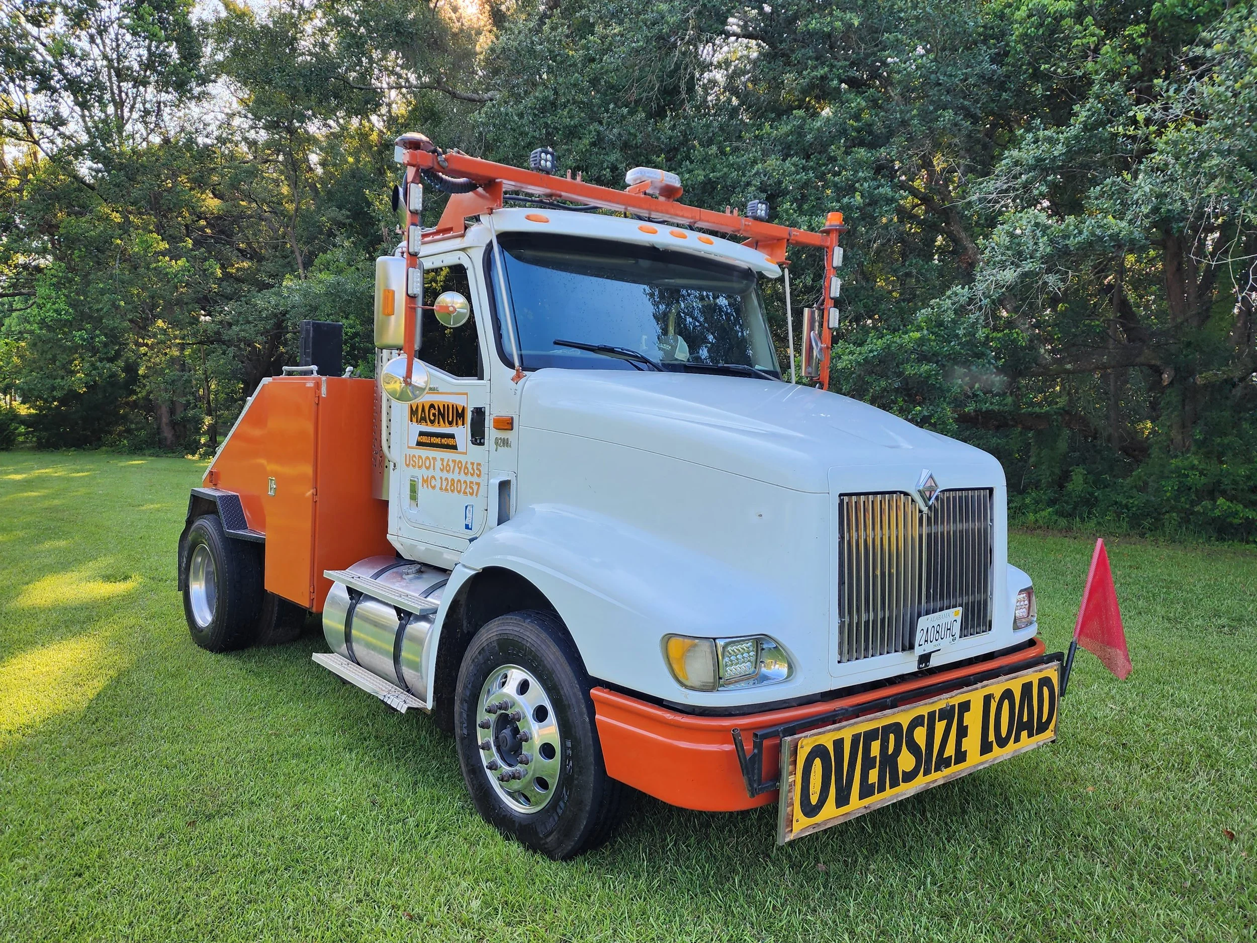 A white tow truck with orange accents and a yellow oversized load sign parked on a grassy area surrounded by trees.