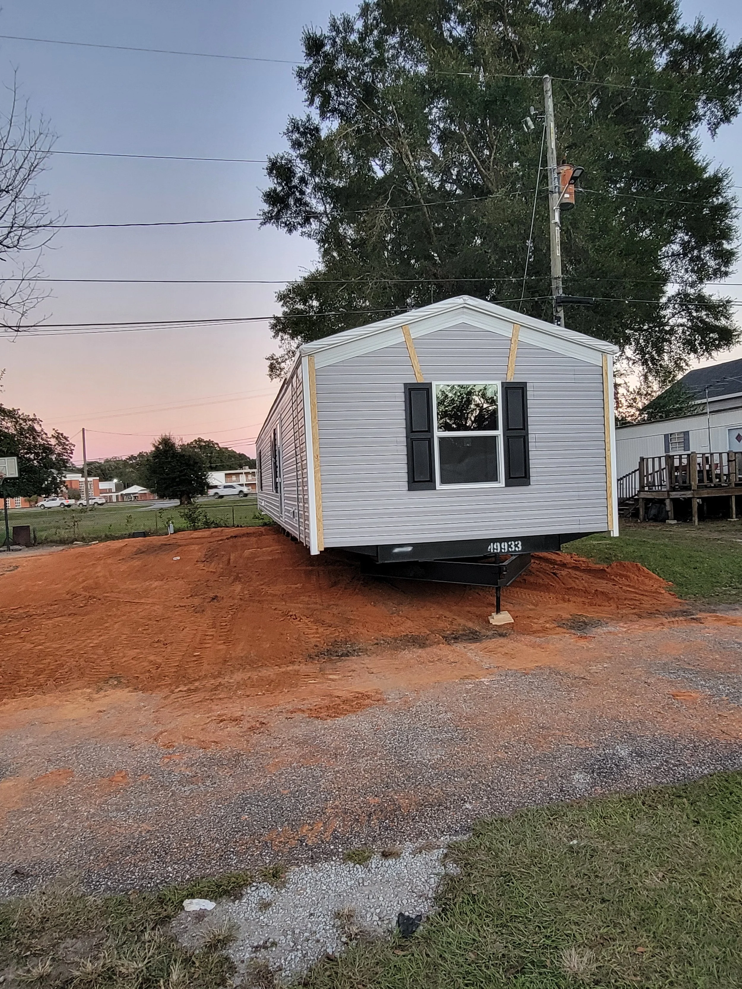 A small white house with black shutters, sitting on a trailer, in a yard with reddish dirt and patches of grass. The background includes trees, power lines, and nearby houses, during sunset or dusk.