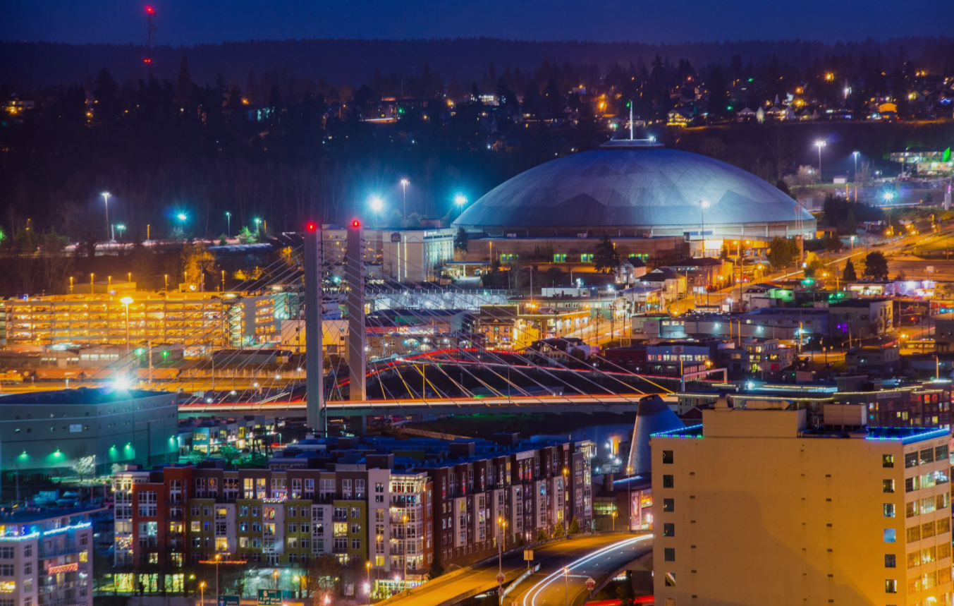 Nighttime cityscape featuring an illuminated cable-stayed bridge, a large domed sports arena, and surrounding buildings with colorful lights.