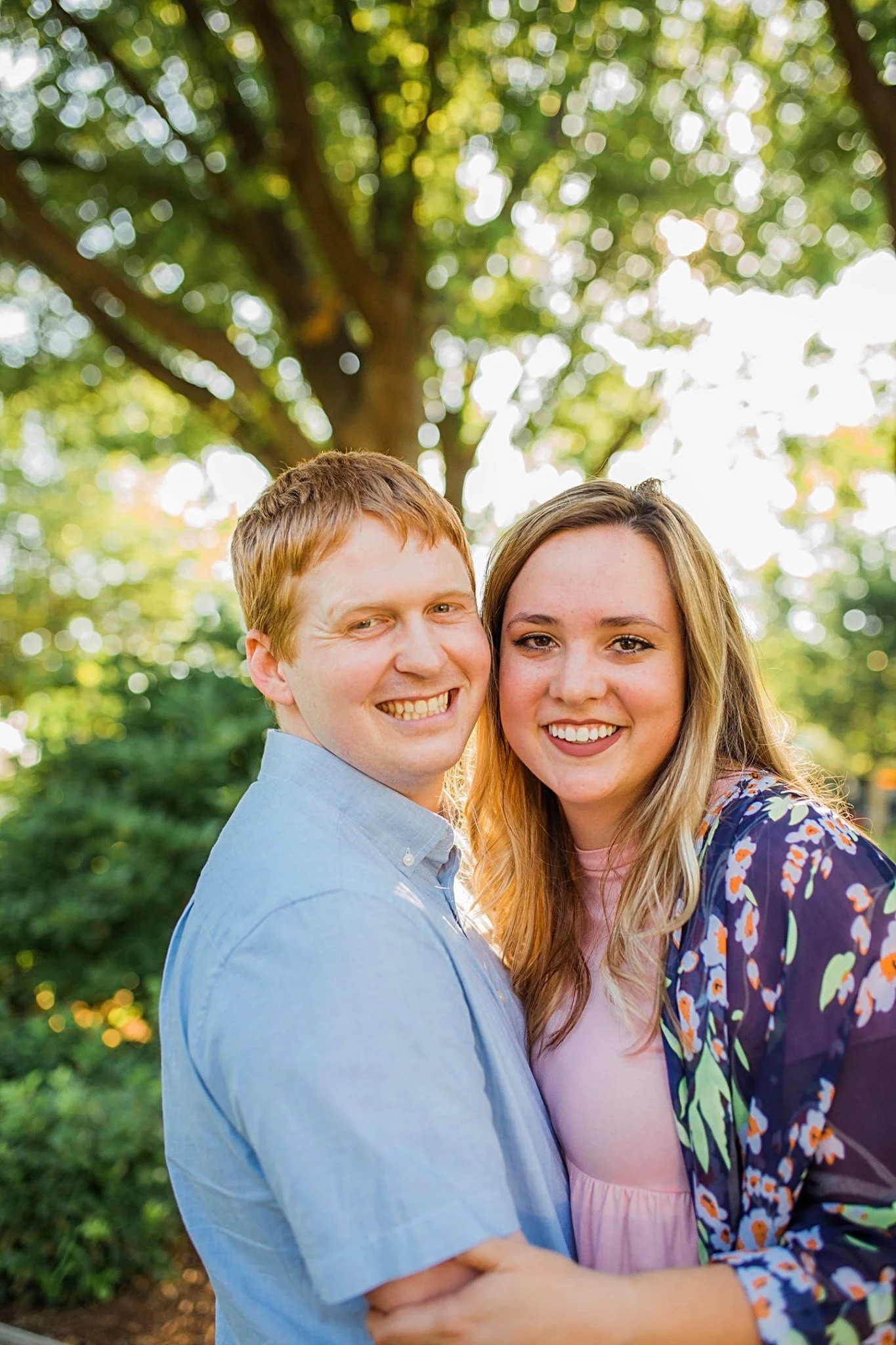 A happy couple is standing outdoors in front of a large tree, smiling and embracing each other. The man has red hair and is wearing a light blue shirt. The woman has long blonde hair and is wearing a pink top with a floral jacket.