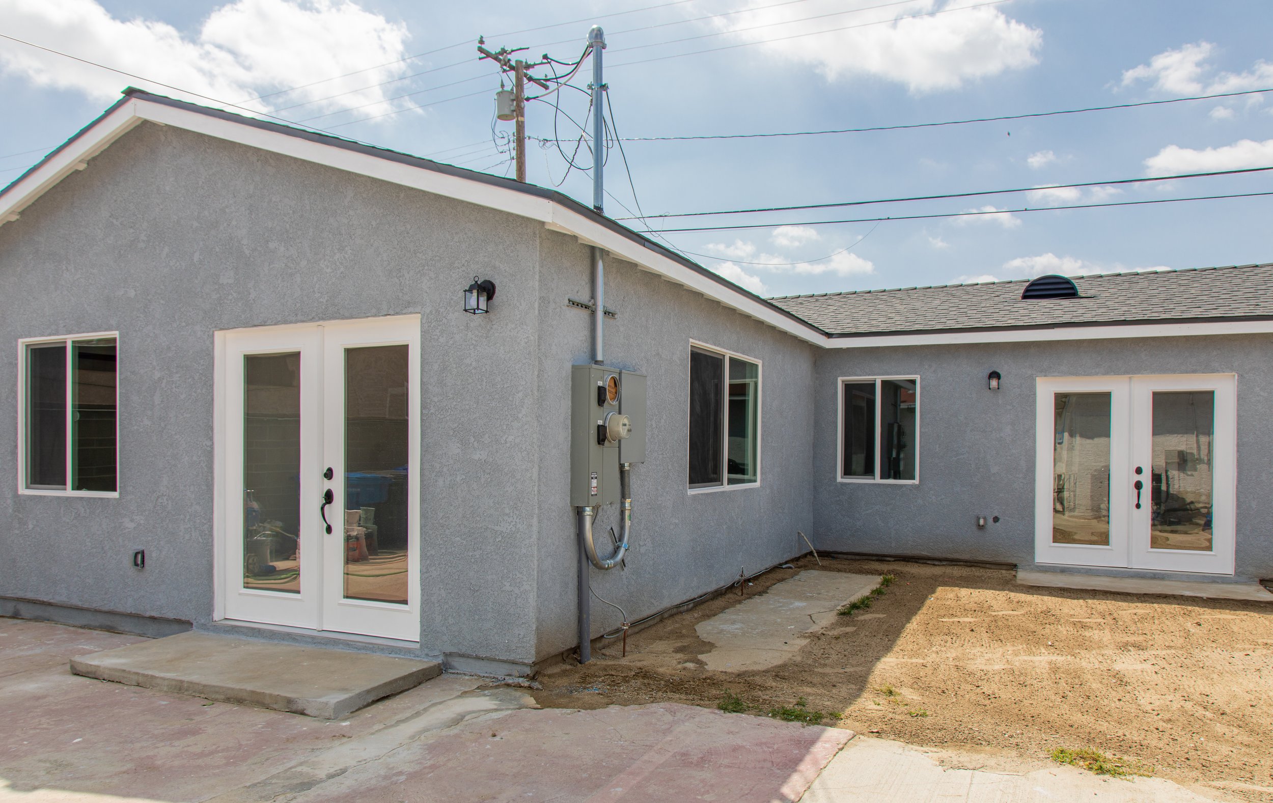 Back view of a newly constructed house with gray stucco exterior, white-framed glass doors, windows, and an electrical meter on the wall, with a dirt yard and concrete pathway.