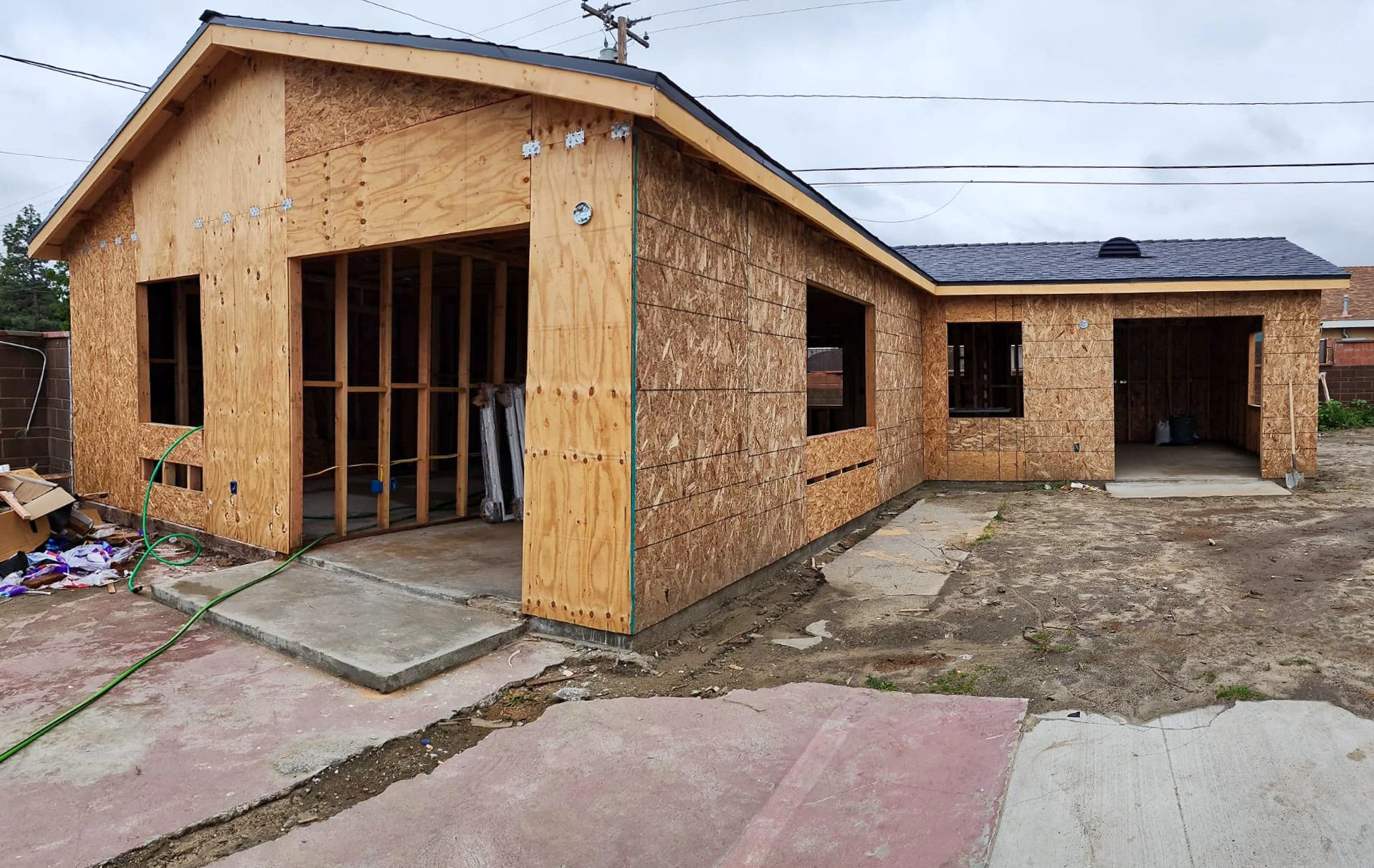 A house under construction with wooden framing and sheathing. The front features an open garage and window openings. The surrounding ground is uneven with construction debris and a green hose lying on a concrete slab.