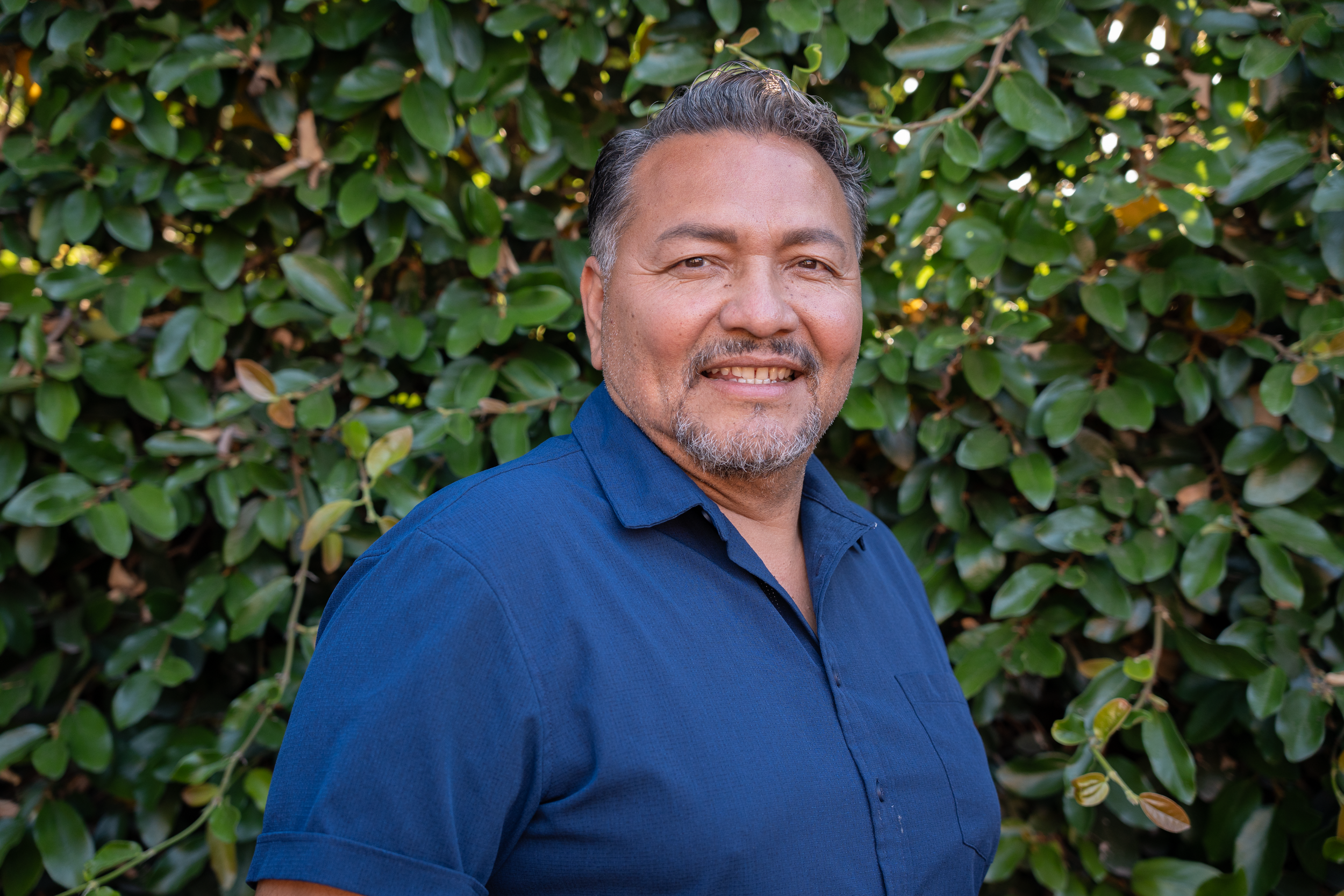 A middle-aged man with gray hair and a short beard, wearing a blue shirt, smiling at the camera in front of a green leafy hedge.