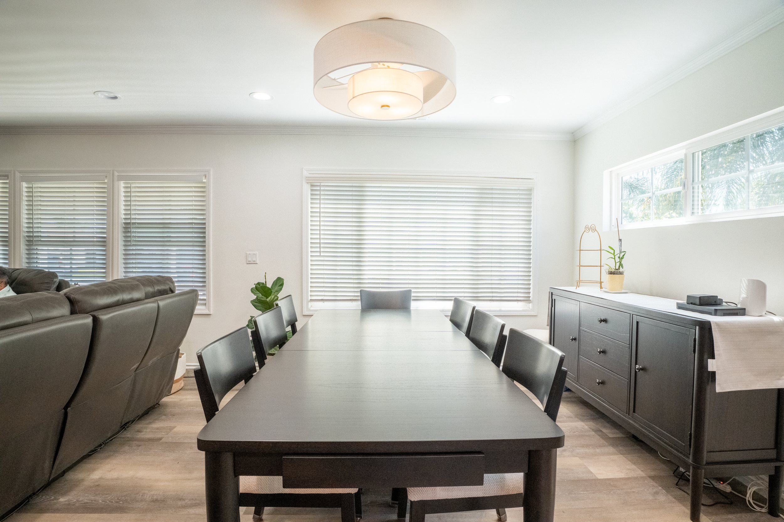 A modern dining room with a large gray table, black chairs, and a gray sideboard. The room has white walls, large windows with blinds, a white ceiling light, and some houseplants.