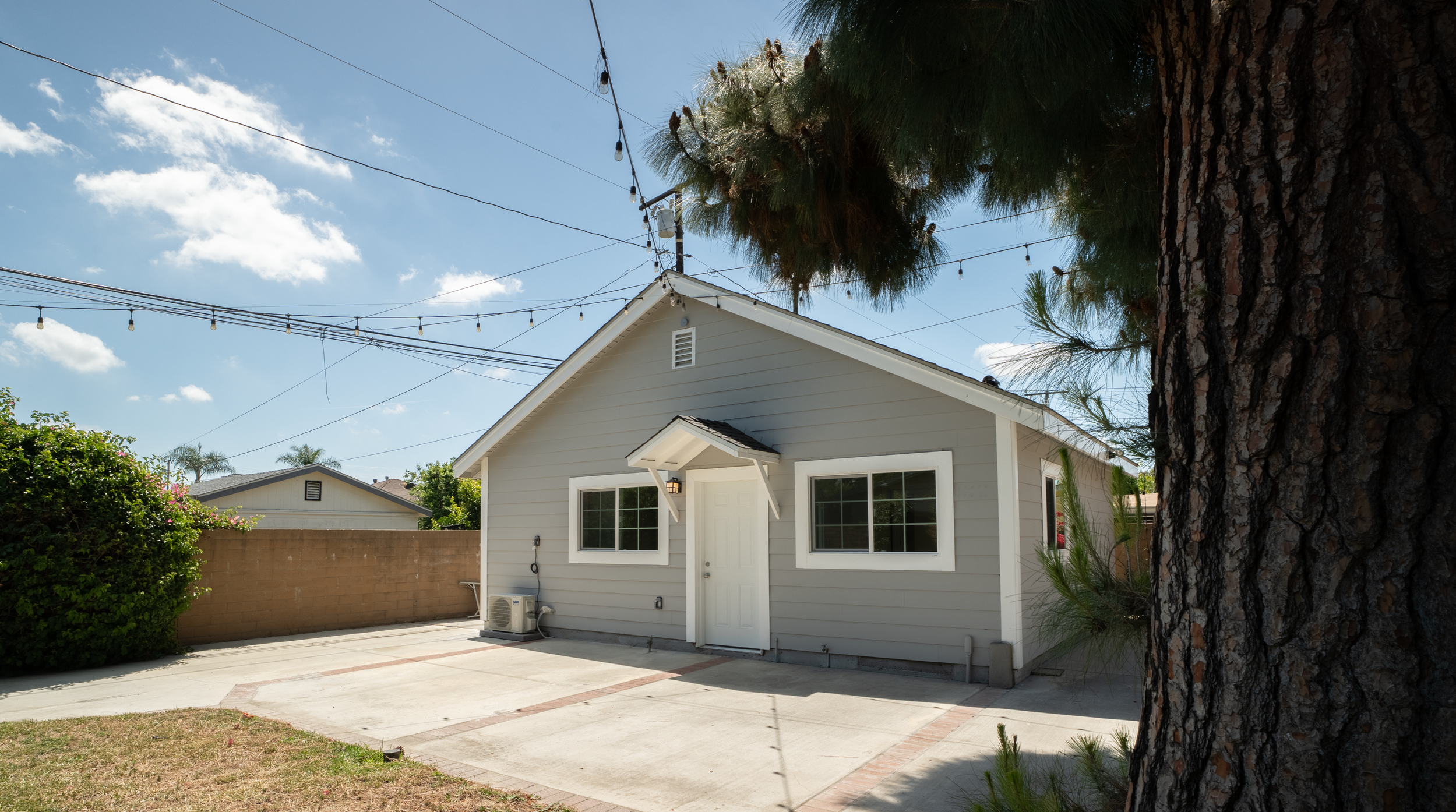 A small white house with a front door and large windows on a sunny day, surrounded by a backyard with a concrete patio, green bushes, and tall trees with overhead string lights.