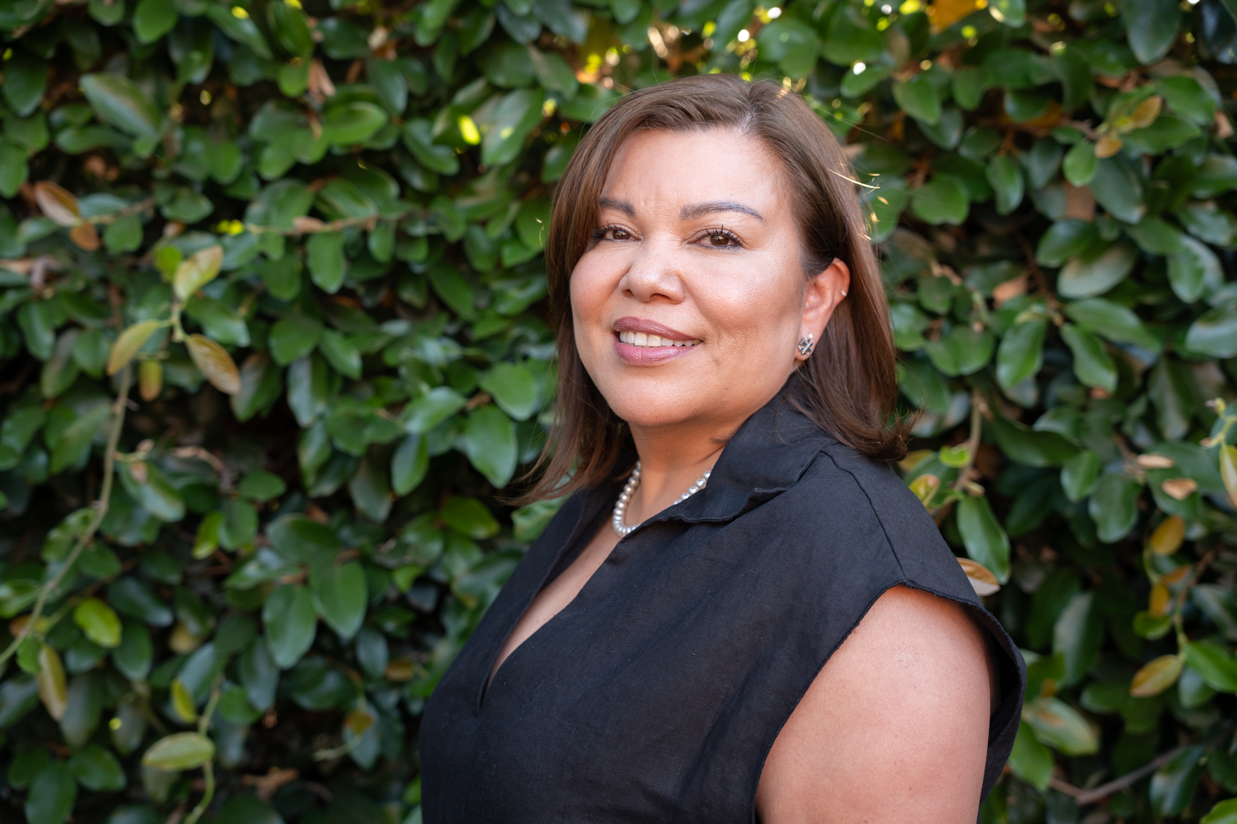 A woman with shoulder-length brown hair wearing a black sleeveless top, pearl necklace, and earrings, standing in front of green leafy bushes.