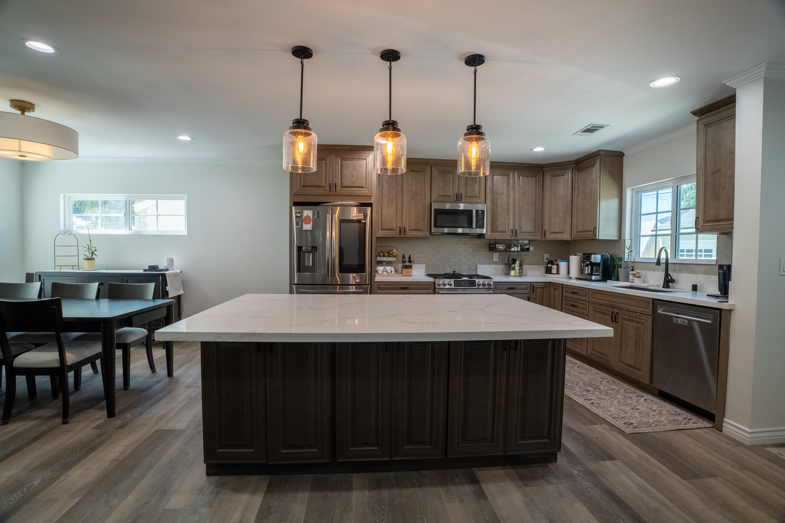 A modern kitchen with wooden cabinets, stainless steel appliances, a large white island, pendant lights hanging above, and a dining area with a table and chairs.
