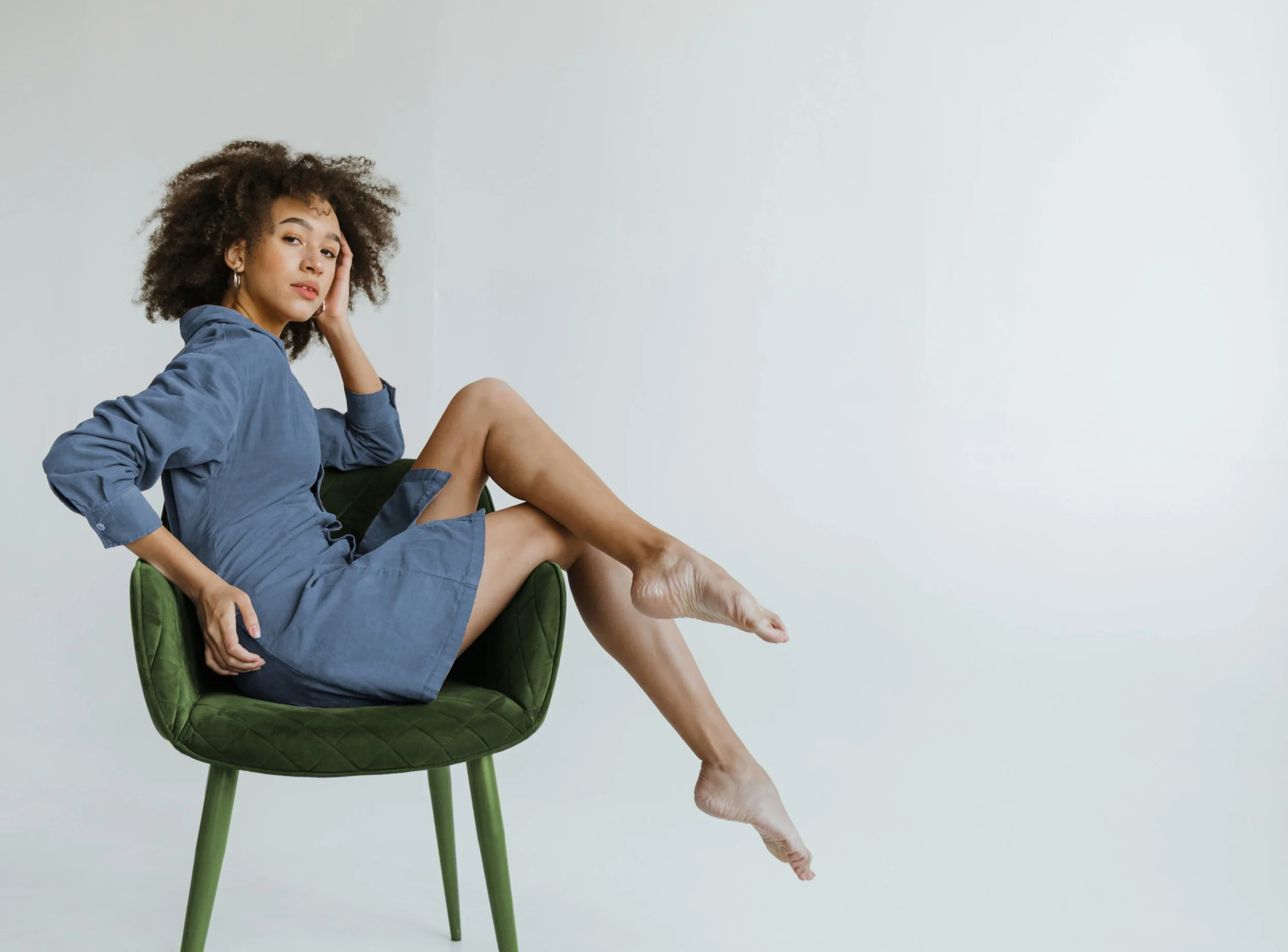 A woman with curly hair wearing a blue hoodie sitting on a green chair against a plain light gray background.