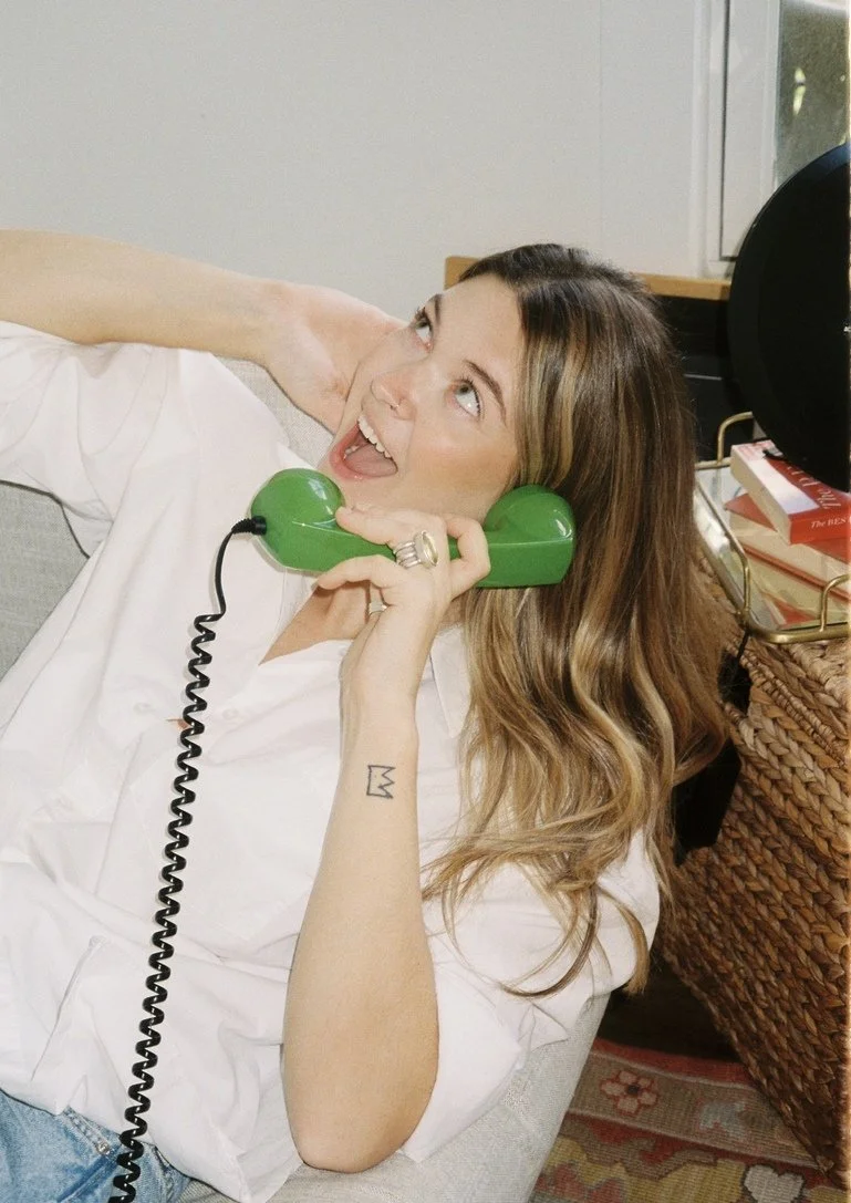 Woman with long wavy hair talking on a bright green retro telephone, smiling and looking upwards, sitting on a beige couch in a cozy room with books and a speaker nearby.
