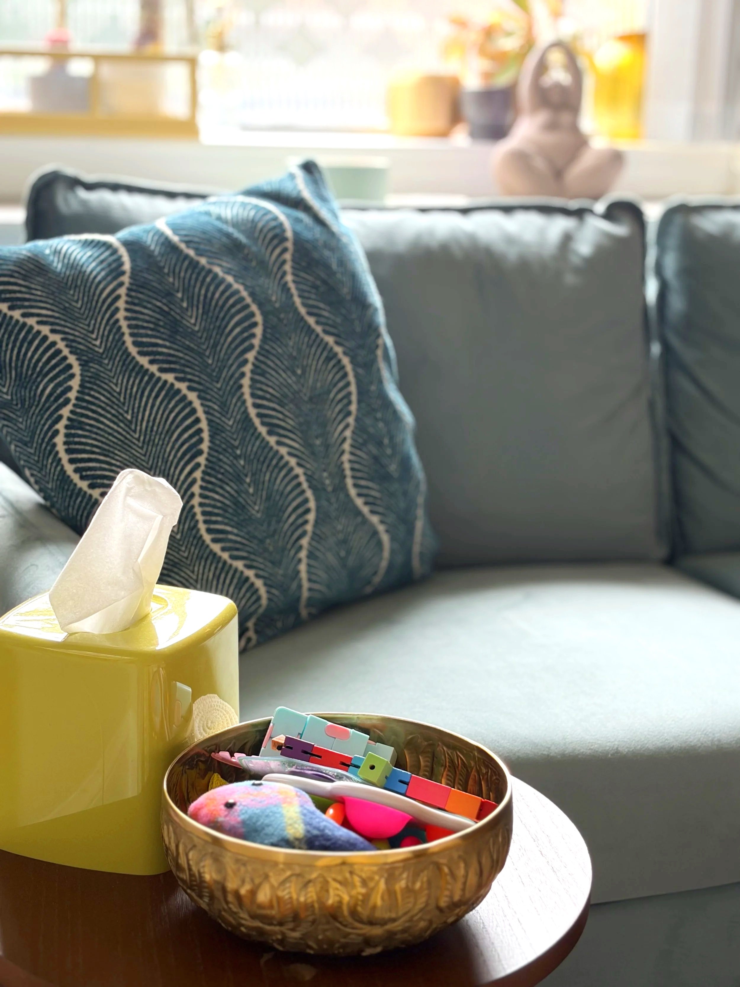 A yellow tissue box and a woven basket filled with colorful items on a wooden table near a gray couch in a bright room with a window.