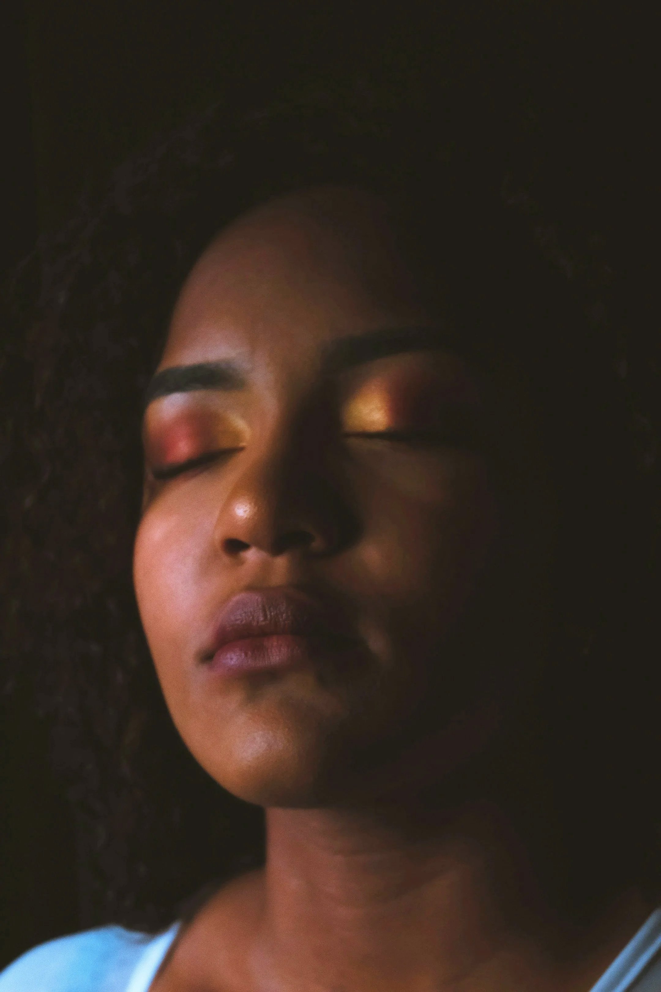 Close-up of a woman's face with her eyes closed, dark curly hair, and wearing a white top.