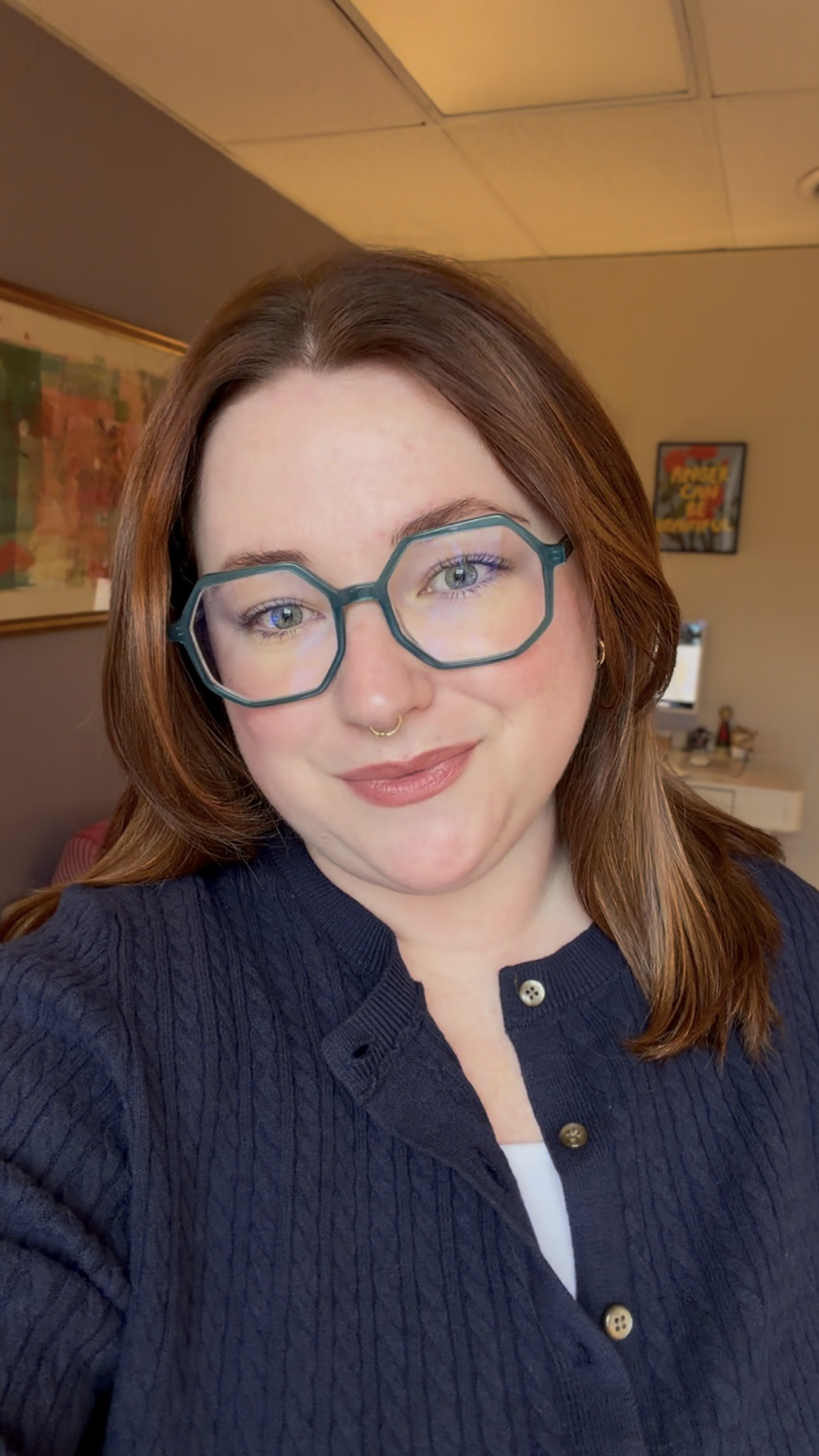A woman with shoulder-length reddish-brown hair, wearing large hexagonal glasses, a nose ring, and a navy blue button-up sweater, smiling at the camera in an indoor setting.