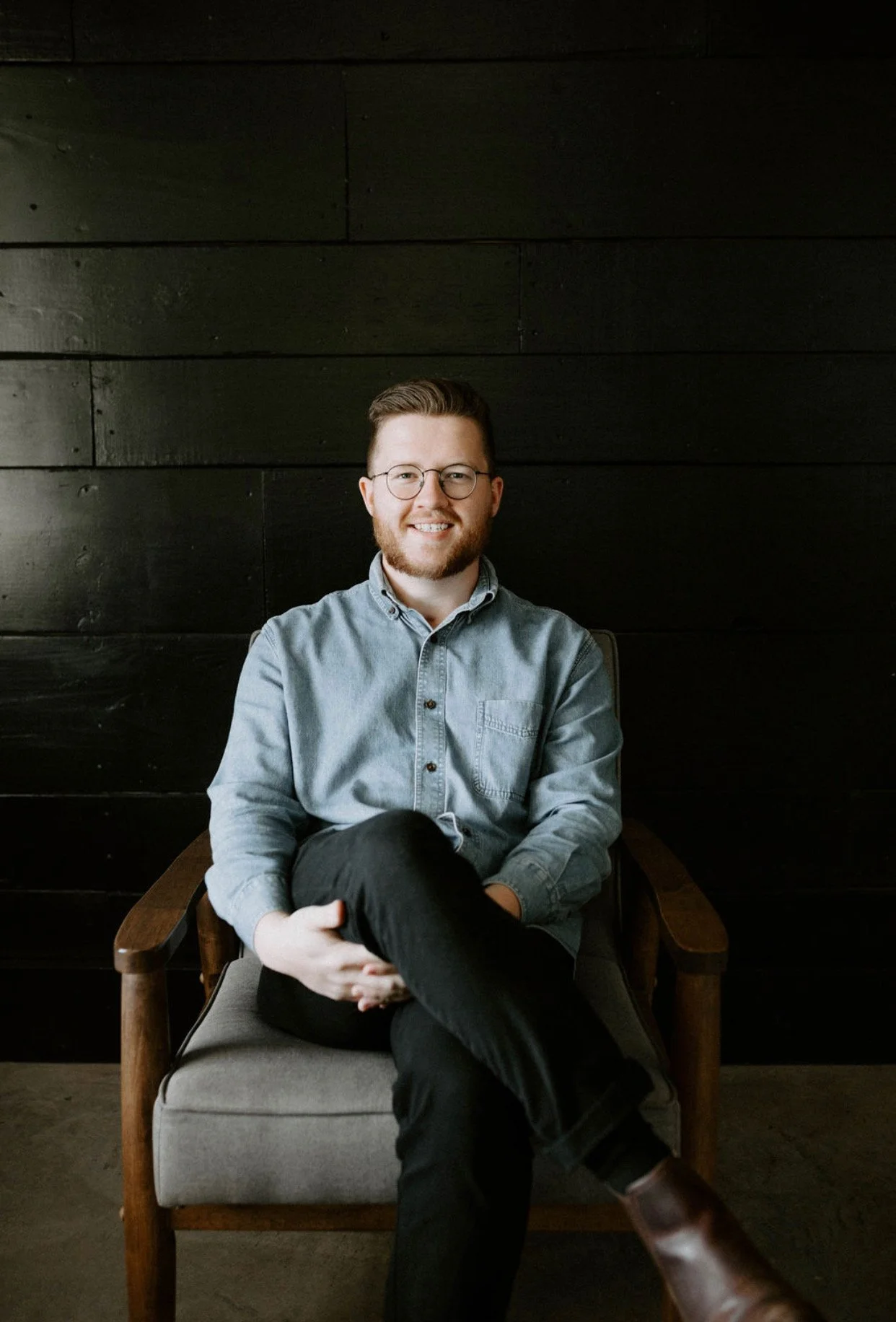 A man with glasses, a beard, and short hair is sitting in a wooden chair with dark fabric cushions, smiling, wearing a light blue button-up shirt and black pants, against a dark wooden wall background.