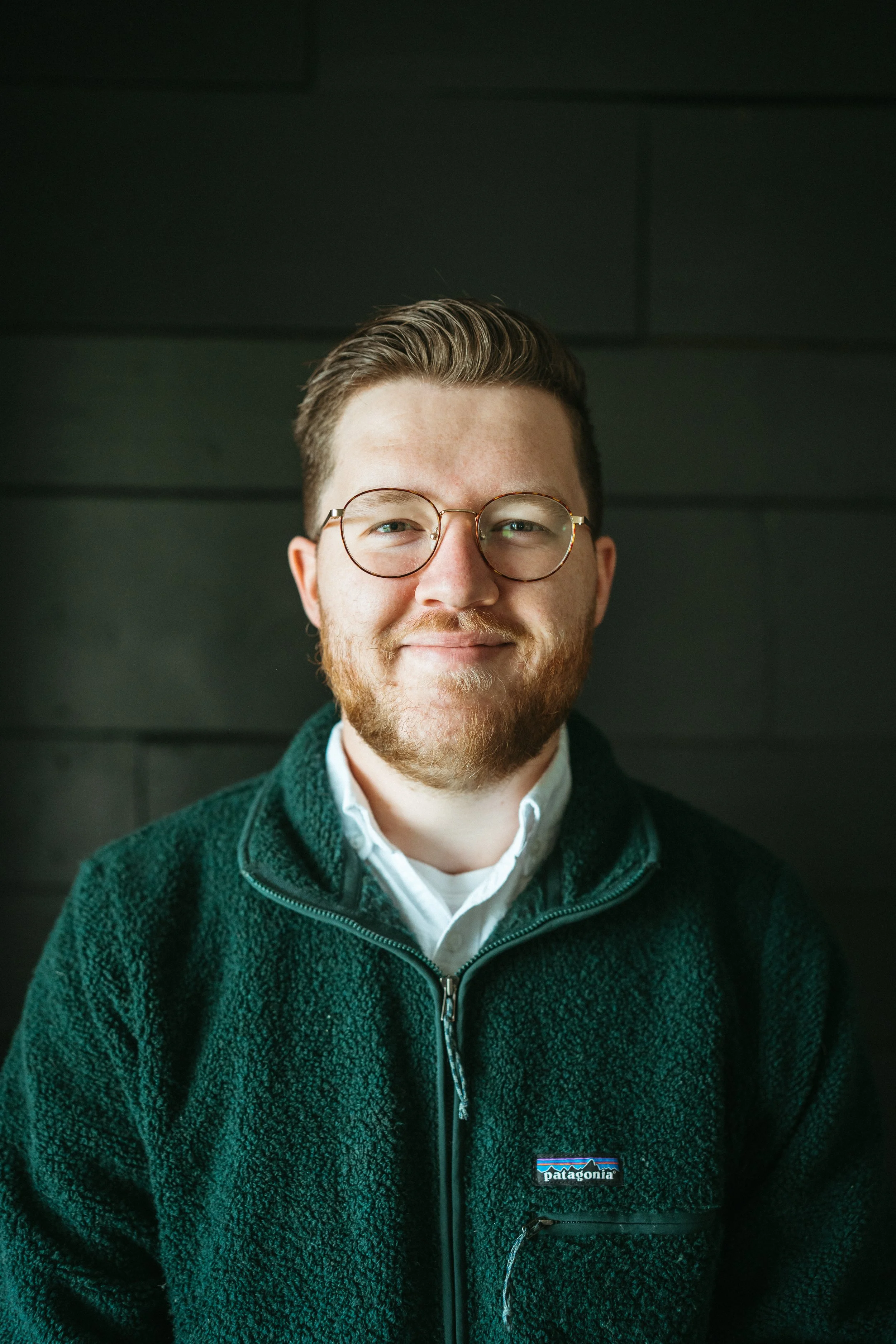 A young man with glasses, a beard, and short hair, smiling, wearing a white shirt and green Patagonia fleece jacket, sitting against a dark background.