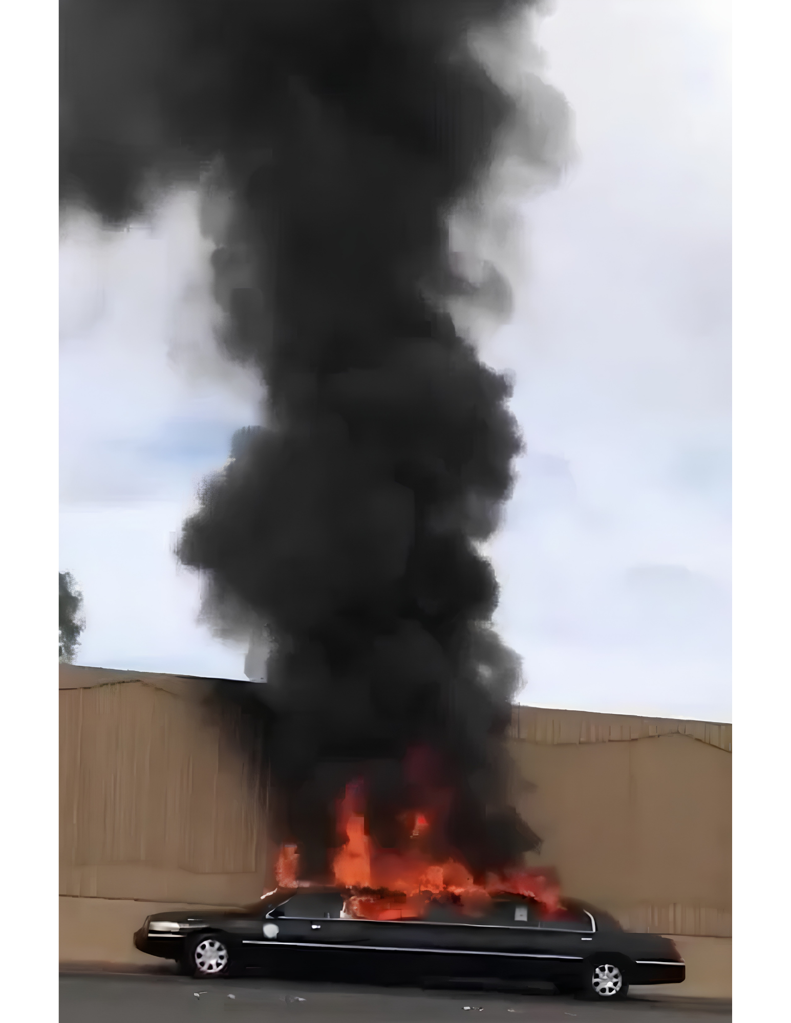 A black car is parked on the street in front of a fire with thick black smoke billowing into the sky. Flames are visible near the back of the fire, with the smoke rising high.