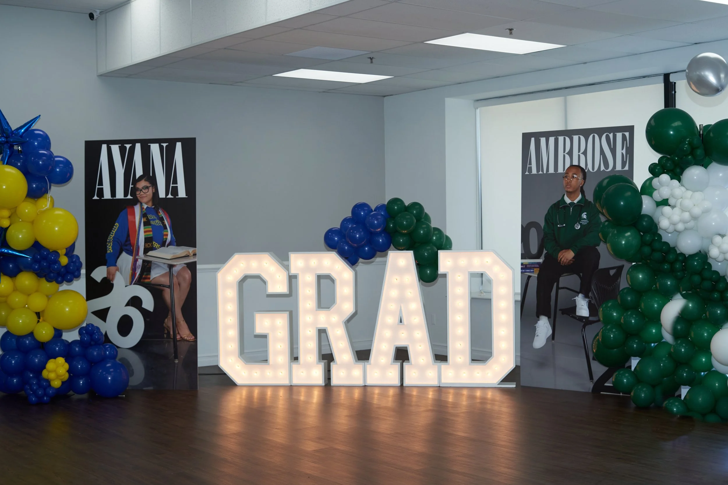 An indoor celebration scene with large illuminated letters spelling 'GRAD' in front of a wall decorated with balloon arrangements in blue, yellow, green, and white. Two individuals are seated on chairs on either side of the letters, each with a poster of a graduate behind them. The person on the left is a smiling woman wearing glasses, a blue jacket, and a rainbow sash, reading a book. The person on the right is a young man in a green jacket with a stethoscope, looking to the side.