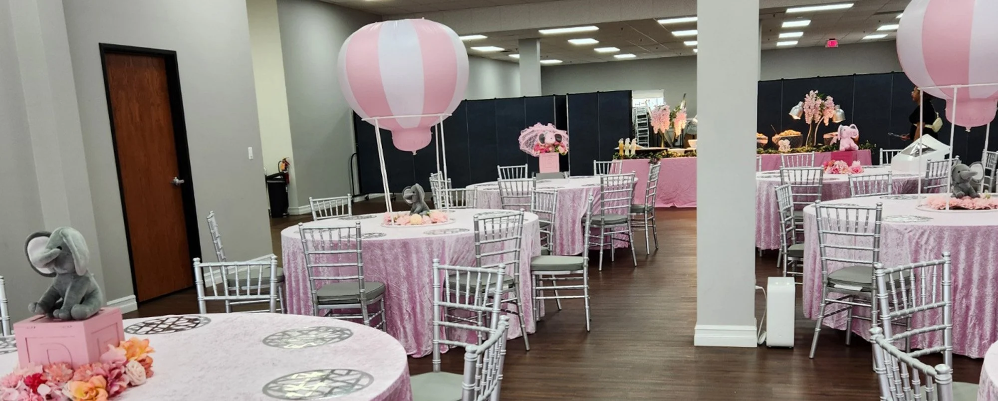 Banquet hall decorated for a party with pink theme including tables with pink tablecloths, large pink paper lanterns, flower arrangements, plush elephants, and pink decorative items.