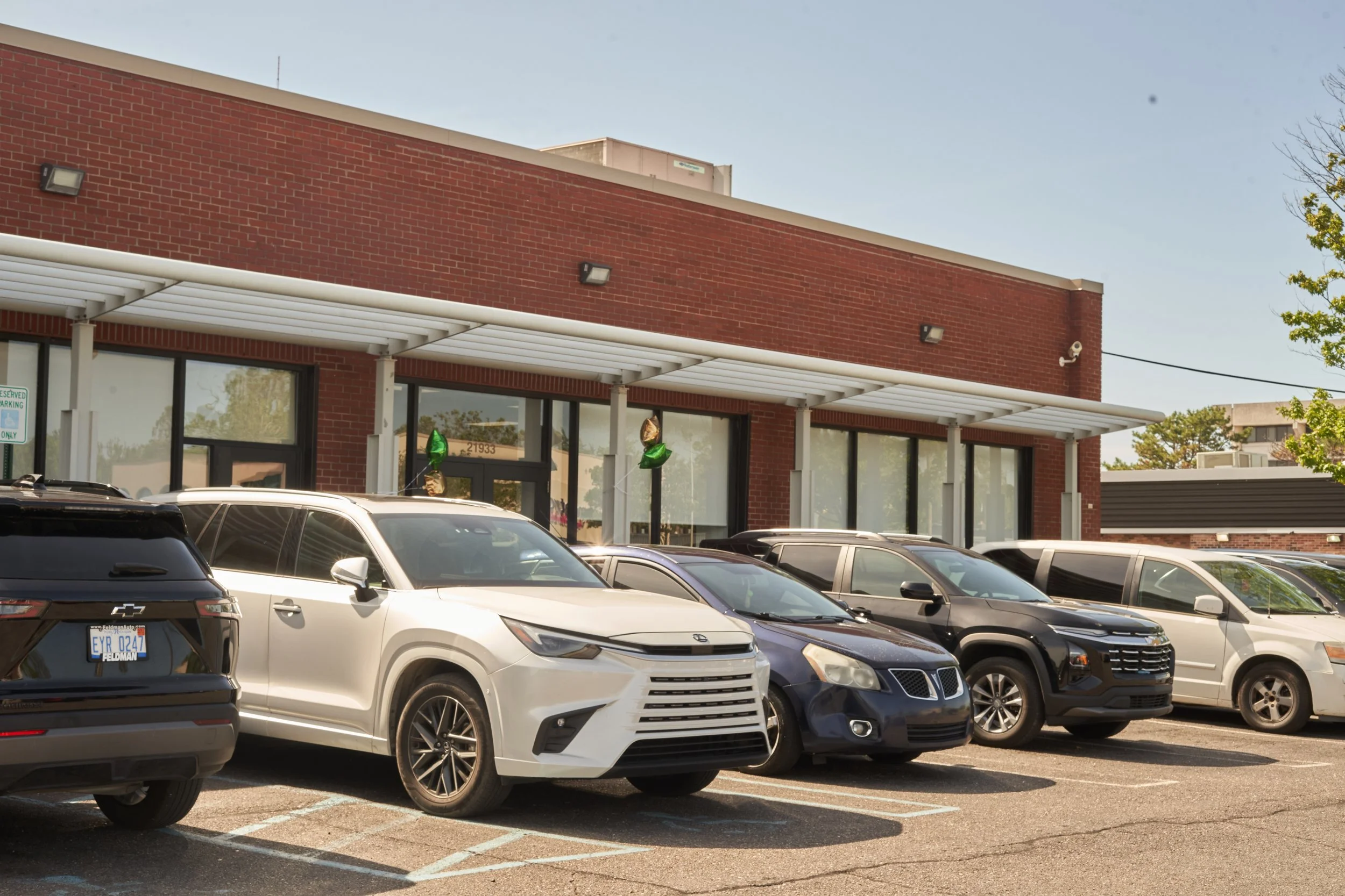 A parking lot filled with various modern cars in front of a red brick building with large glass windows and a white awning, under a clear blue sky.