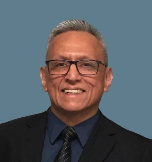 A middle-aged man with short gray hair, glasses, and light skin, smiling and wearing a dark suit, navy shirt, and a dark striped tie against a solid blue background.