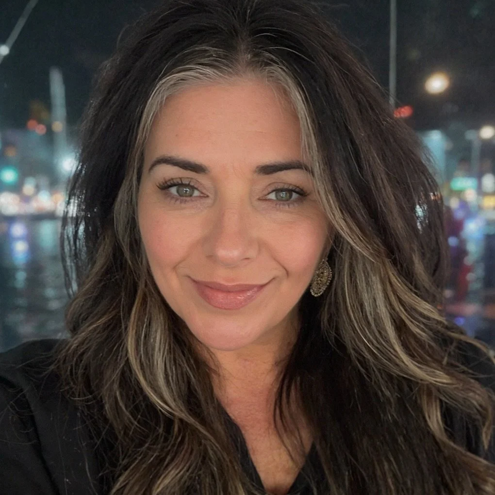 A woman with long, wavy brown hair with blonde highlights smiling at the camera, with city lights and blurred cityscape in the background at night.