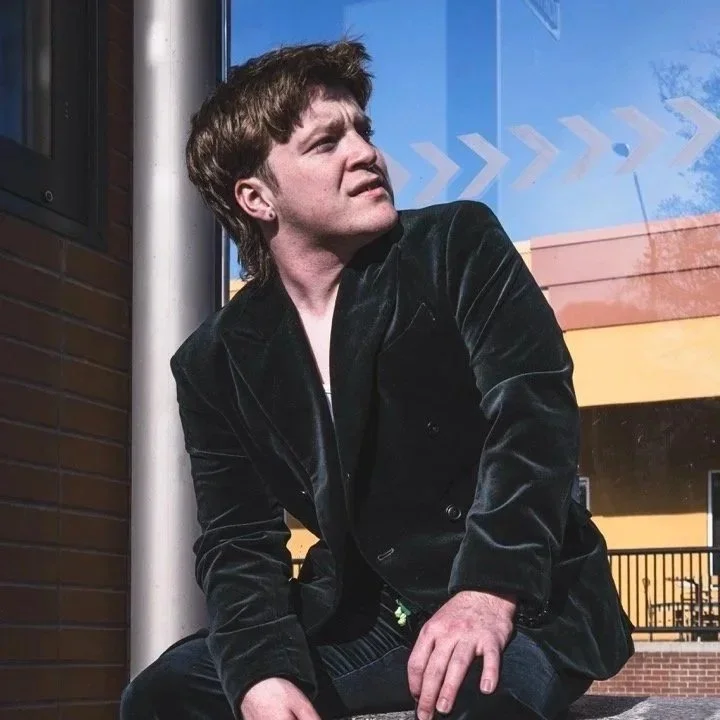 A young man with dark hair sitting outside near a brick wall and a large window with blue sky and clouds reflected in it.