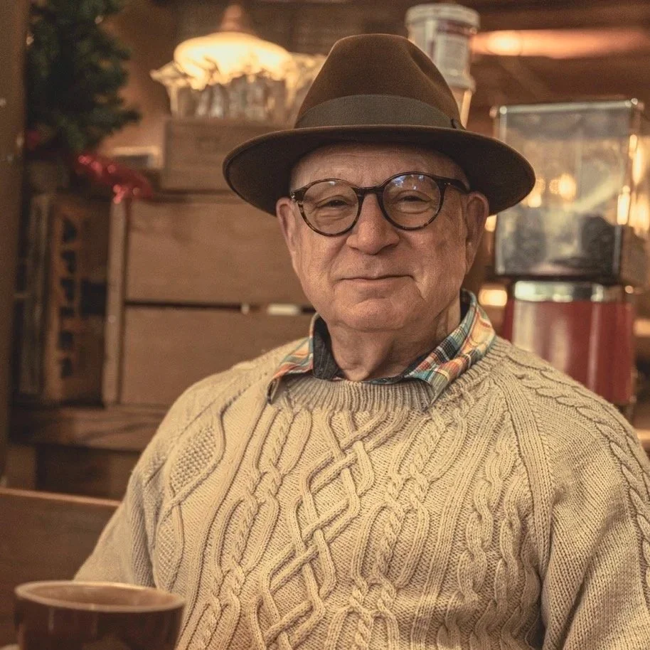 An elderly man wearing glasses, a brown fedora, a plaid shirt, and a beige cable-knit sweater, smiling indoors with warm lighting and rustic wooden decor.
