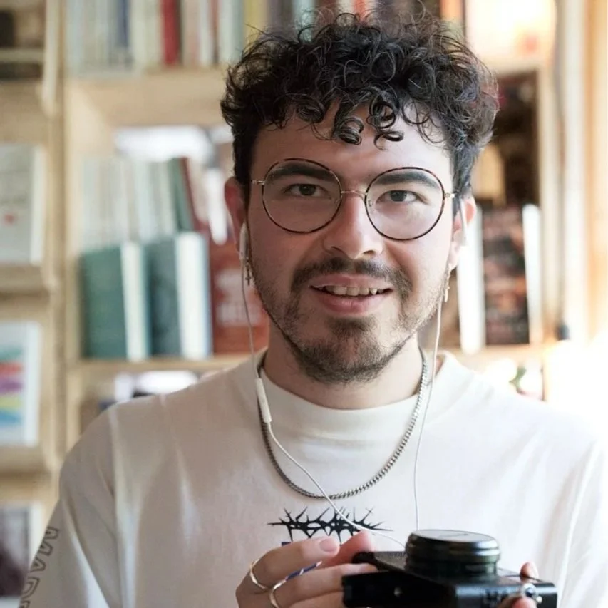 A young man with curly dark hair, glasses, and a beard, smiling while holding a camera indoors. He is wearing a white shirt with a graphic design and earphones, with bookshelves in the background.