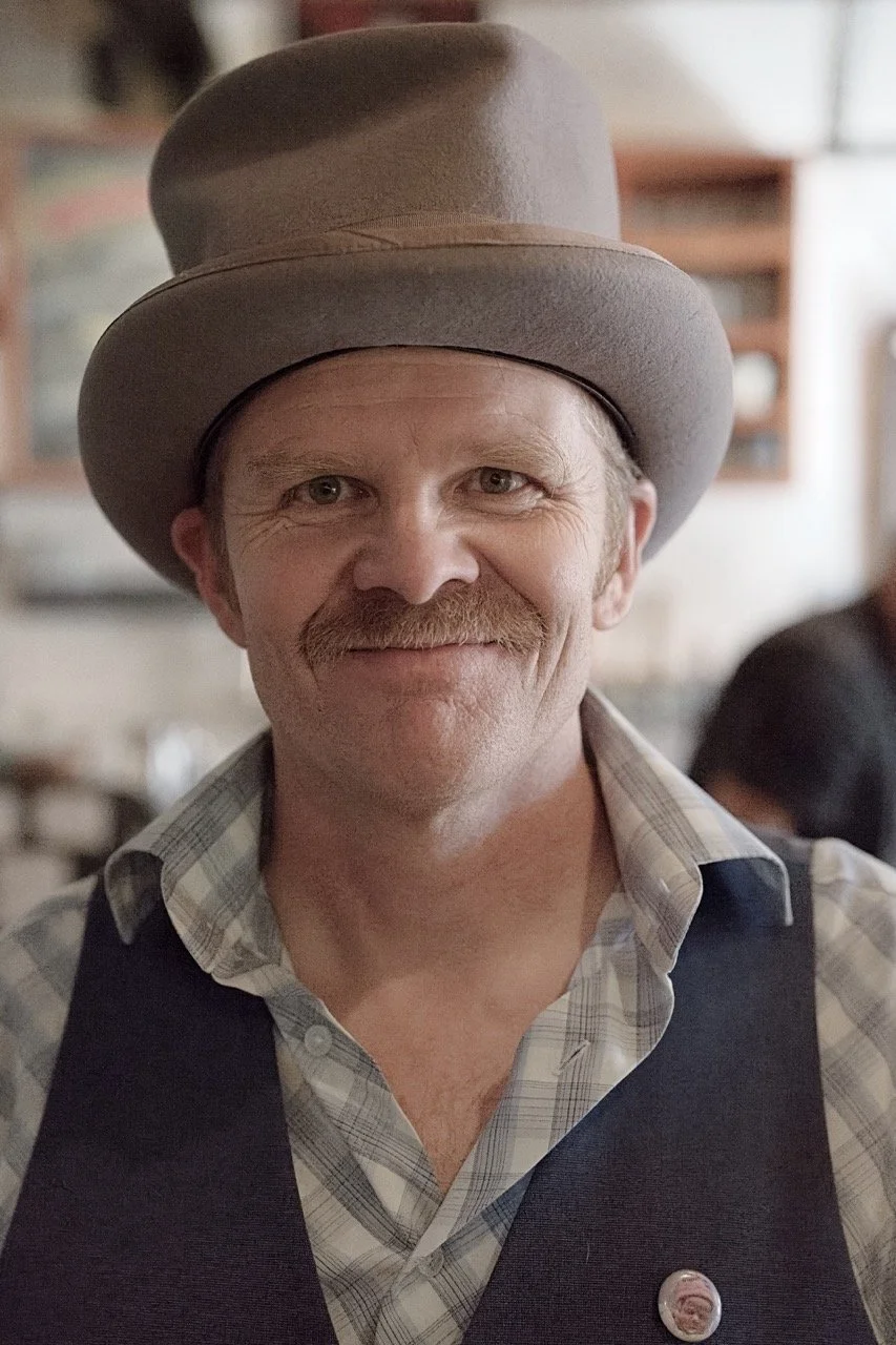 A man with a mustache and light-colored hair wearing a gray top hat, plaid shirt, and dark vest, smiling at the camera in a rustic setting.