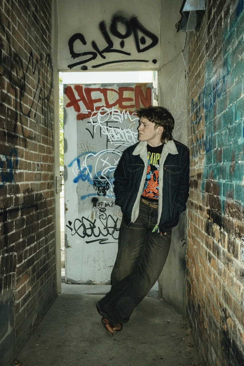A person with short dark hair leaning against a graffiti-covered wall in a narrow alleyway, wearing a black jacket with a shearling collar, a graphic t-shirt, baggy pants, and brown shoes.