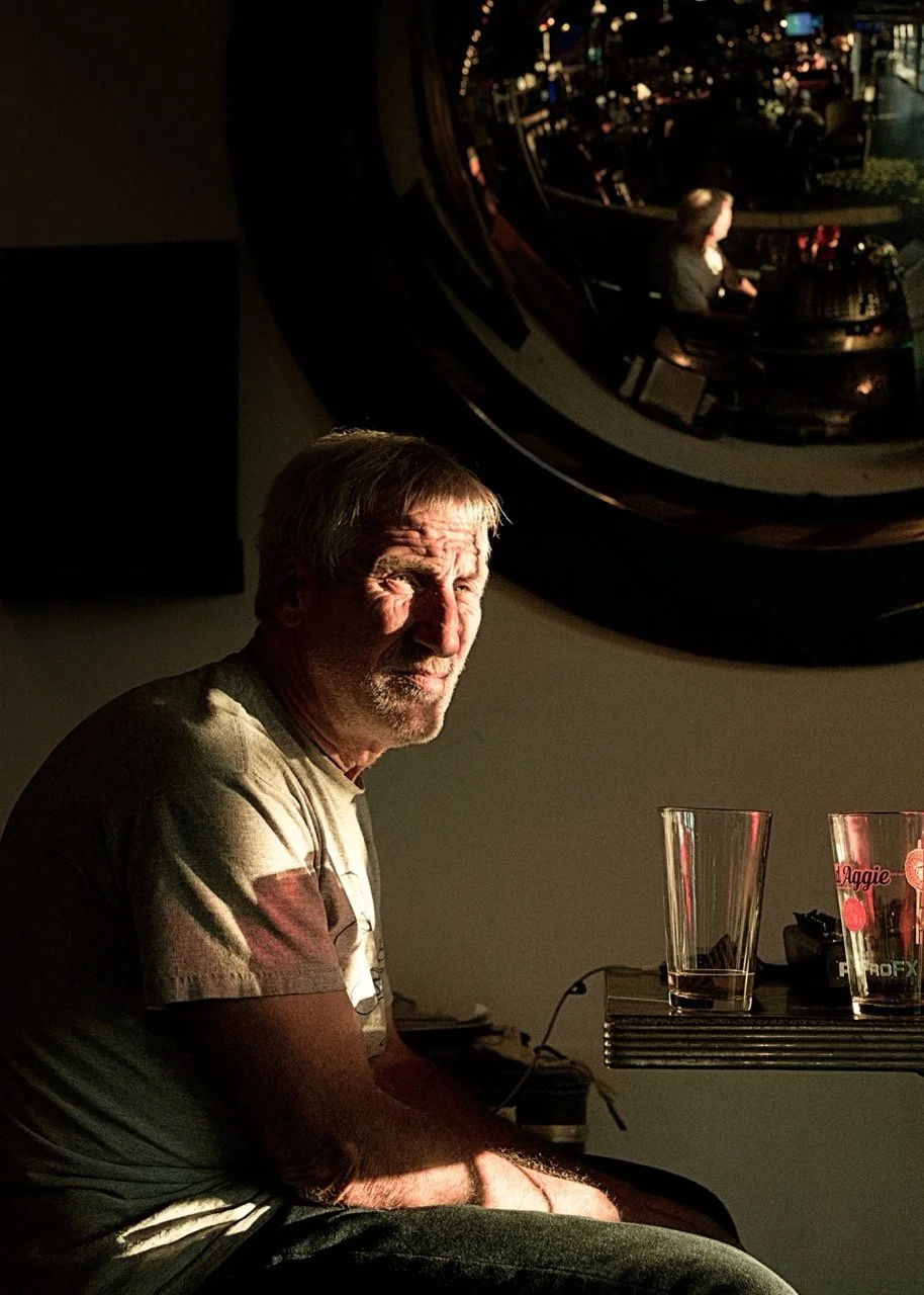 A man sitting indoors with warm sunlight on his face, near a dark wall. A large, round, reflective mirror on the wall shows a woman sitting at a piano in the background. There are glasses and a glass of water on a table beside him.