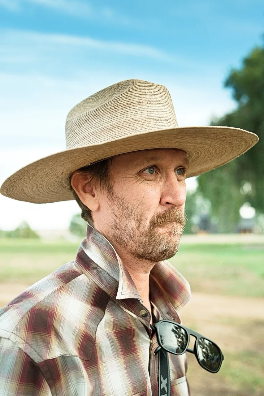 A man wearing a large straw hat, plaid shirt, and sunglasses hanging from his collar, standing outdoors in a rural setting with trees and fields in the background.