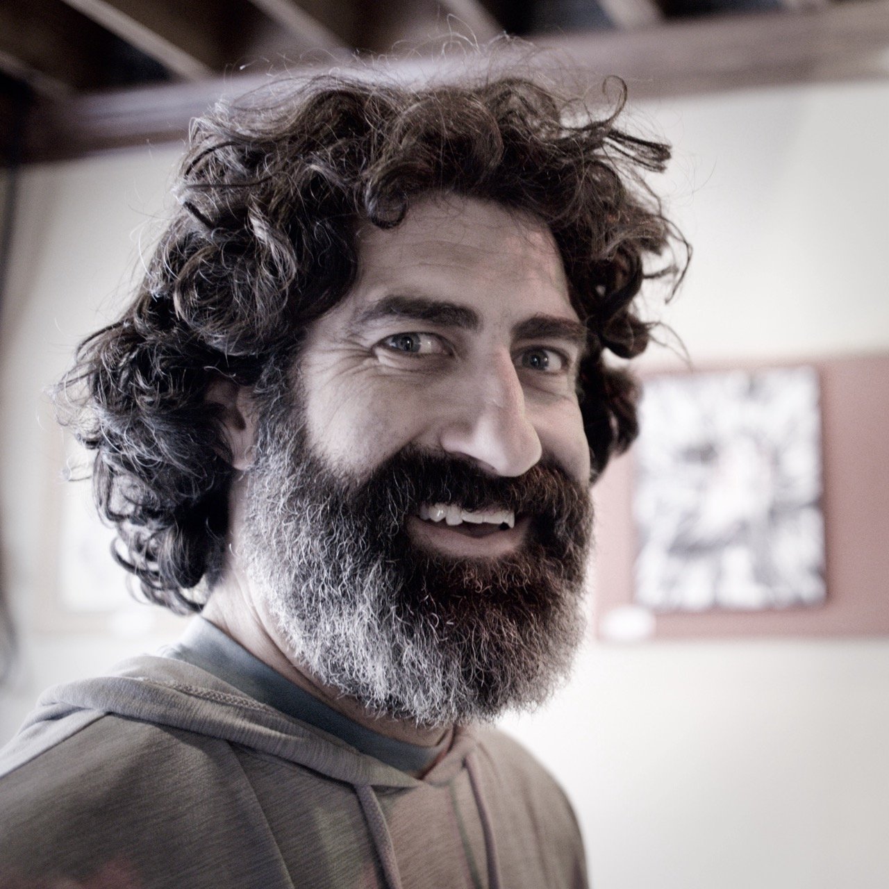 A man with curly hair and a beard smiling in an indoor setting.