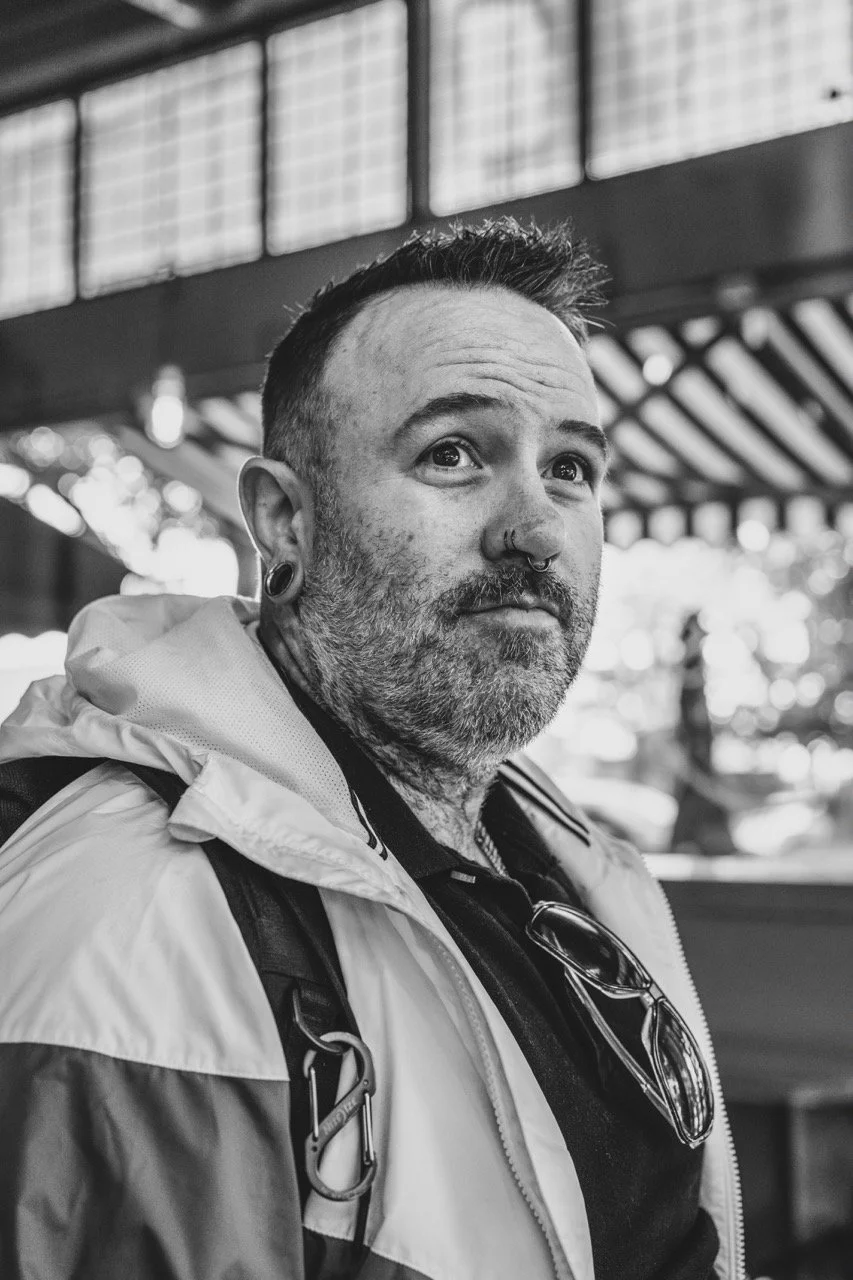 A black and white photo of a bearded man with tattoos, piercings, and a backpack, looking slightly to the side, outdoors with blurred background.
