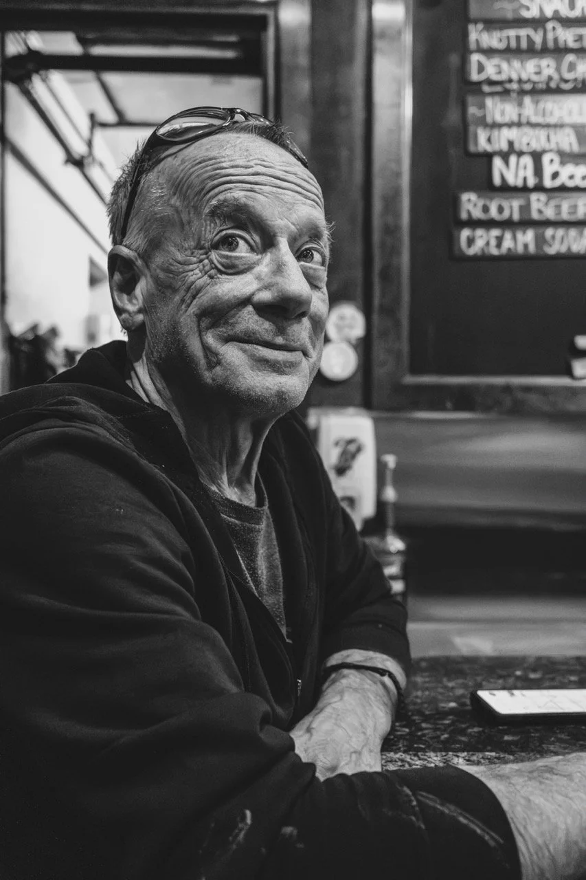 A black-and-white photo of an older man with glasses on his head, sitting at a bar or restaurant counter, smiling at the camera, with a menu board in the background.