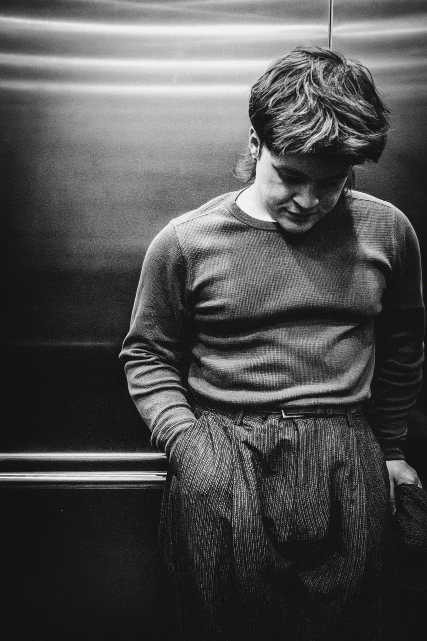 A young man with wavy hair standing in an elevator, looking down with his hands in his pockets, dressed in a long-sleeve shirt and striped pants, in black and white.